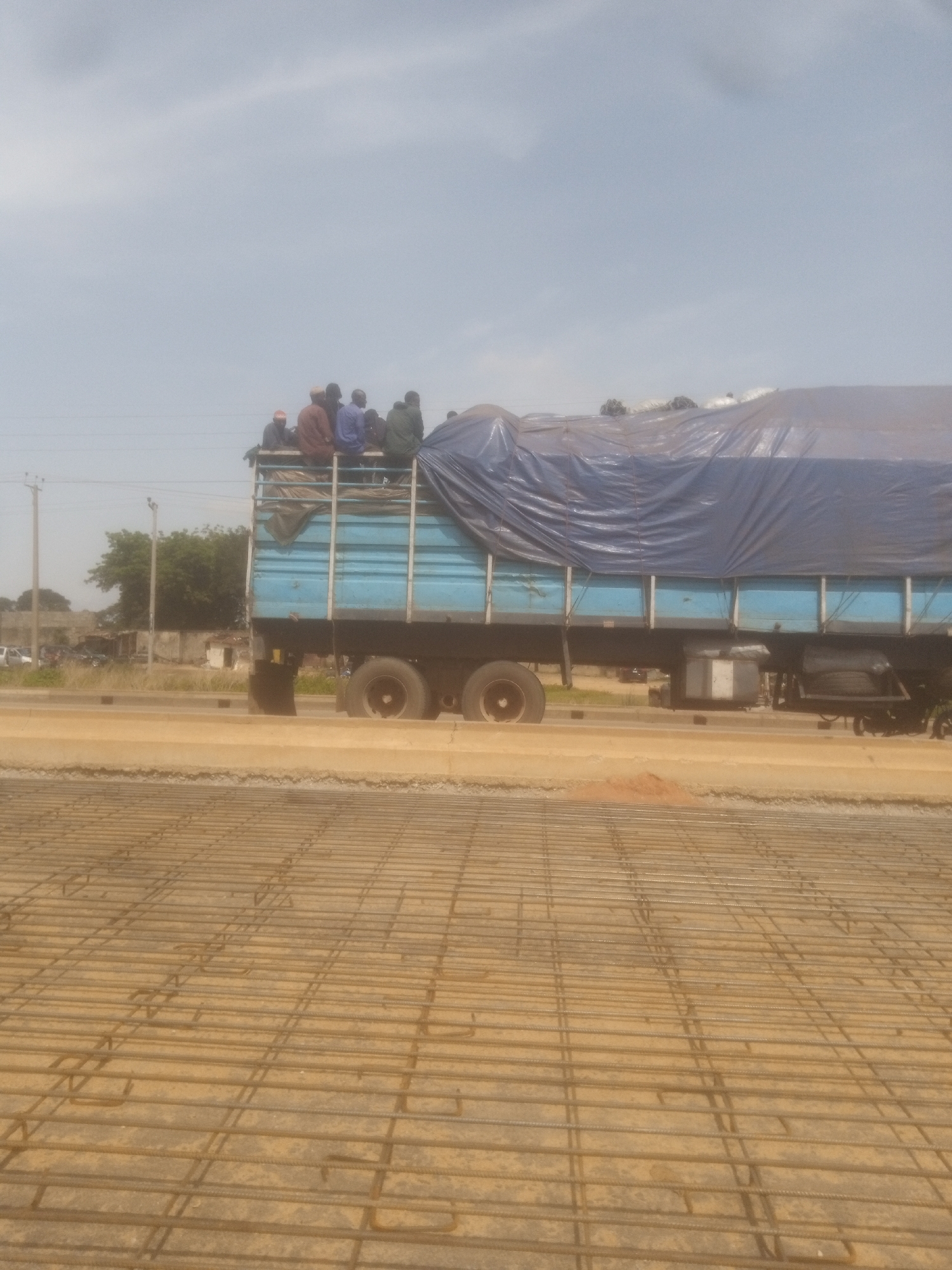 In Nassarawa, Nigeria, a flatbed truck is traveling on a paved road. The truck is loaded with cargo covered by a large blue tarp. On top of the tarp, approximately eight to ten individuals are seated, appearing to be passengers. The truck is a light blue color and has two visible axles with dual rear wheels. To the left of the truck, there are utility poles, trees, and some buildings. In the foreground, a construction site is visible, with a grid of metal rebar laid out on the ground. The sky is overcast with some clouds.
