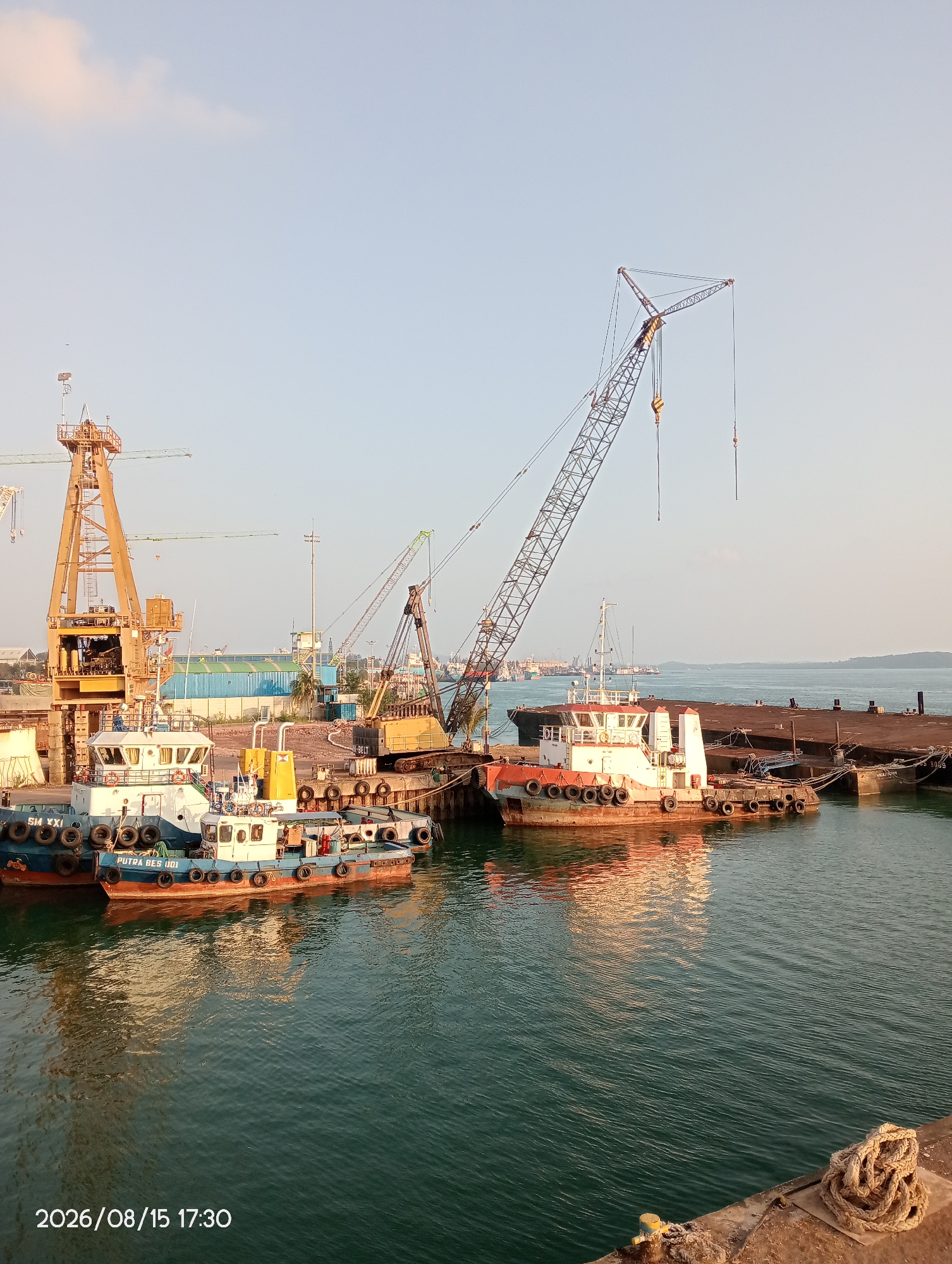 The image shows a harbor in Batam, Indonesia, with several tugboats docked at a pier. A large construction crane is visible in the background, along with other industrial equipment and buildings. The water is calm, reflecting the sky and the vessels. The timestamp on the image indicates it was taken on August 15, 2026, at 17:30.