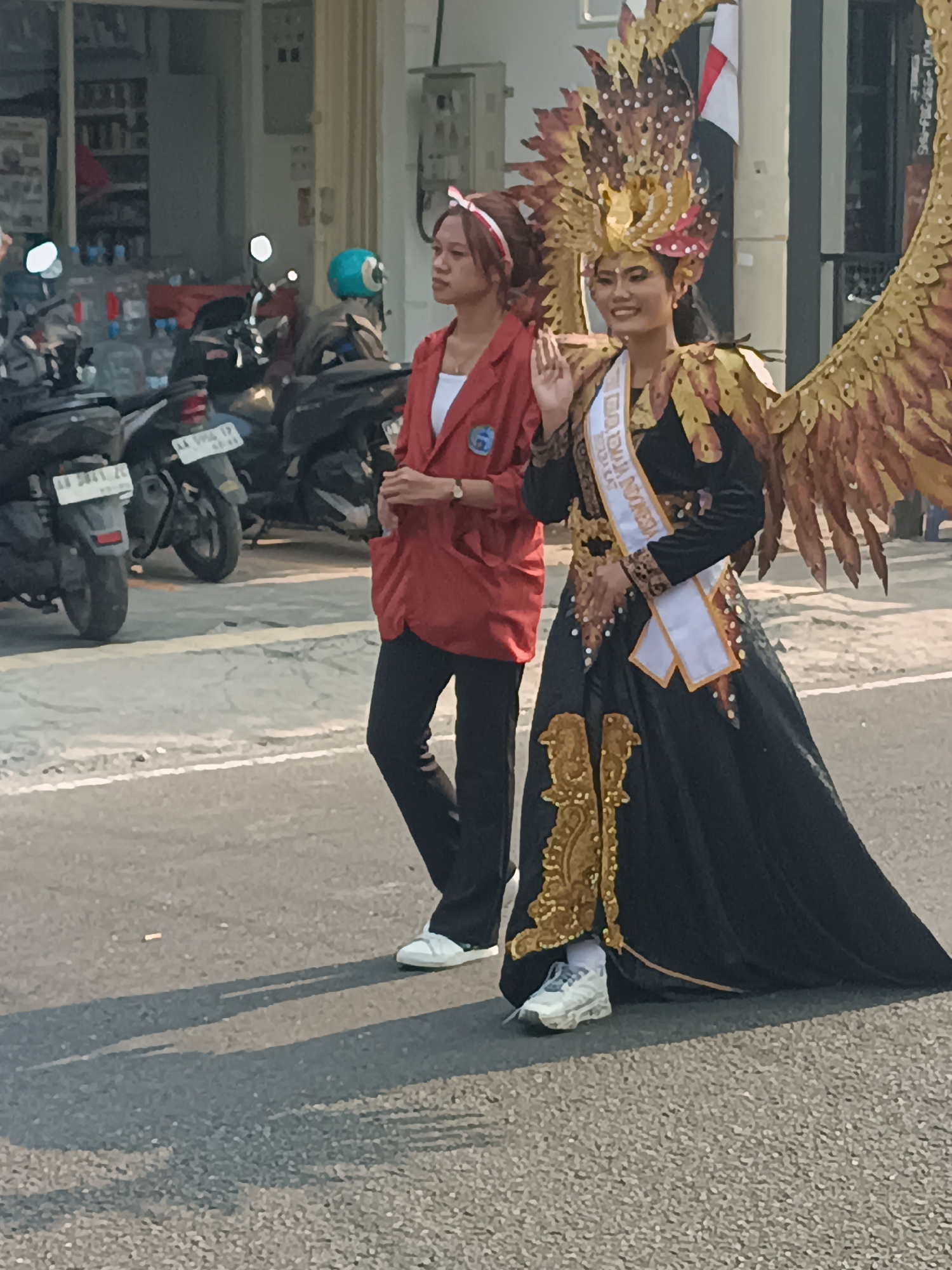 In Sleman, Indonesia, a street scene unfolds with two women, one in a vibrant red blazer and the other adorned in an elaborate golden headdress and black gown. The woman in the red blazer stands on the left, her hand resting on her hip, while the woman in the costume is positioned on the right, her palm raised in a graceful wave. Behind them, a row of motorcycles are parked along the sidewalk, suggesting a busy urban or semi-urban environment. The sun casts long shadows on the asphalt, indicating it is either early morning or late afternoon. The backdrop of the scene features a building with a clean facade, implying a commercial or public space. The image captures a moment of what appears to be a parade or a cultural event, given the festive attire of one of the individuals.