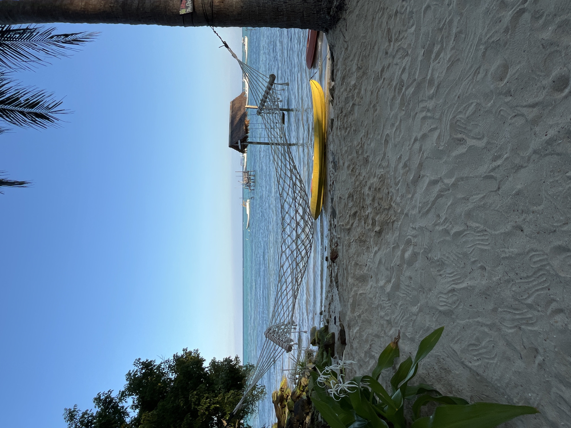 This image depicts a tropical beach scene on a clear day, likely in Legonipah, Indonesia. In the foreground, a sandy beach shows textured patterns from receding waves and footprints. A lush green plant with white flowers grows on the left side. A hammock made of net is suspended between a tree on the left and a palm tree on the right. In the water, a yellow kayak and a red canoe rest on the shore. Further out, a thatched-roof structure stands in the shallow water, with several boats anchored in the distance. The ocean is a calm blue, meeting the clear blue sky at the horizon. The overall atmosphere is one of tranquility and natural beauty.