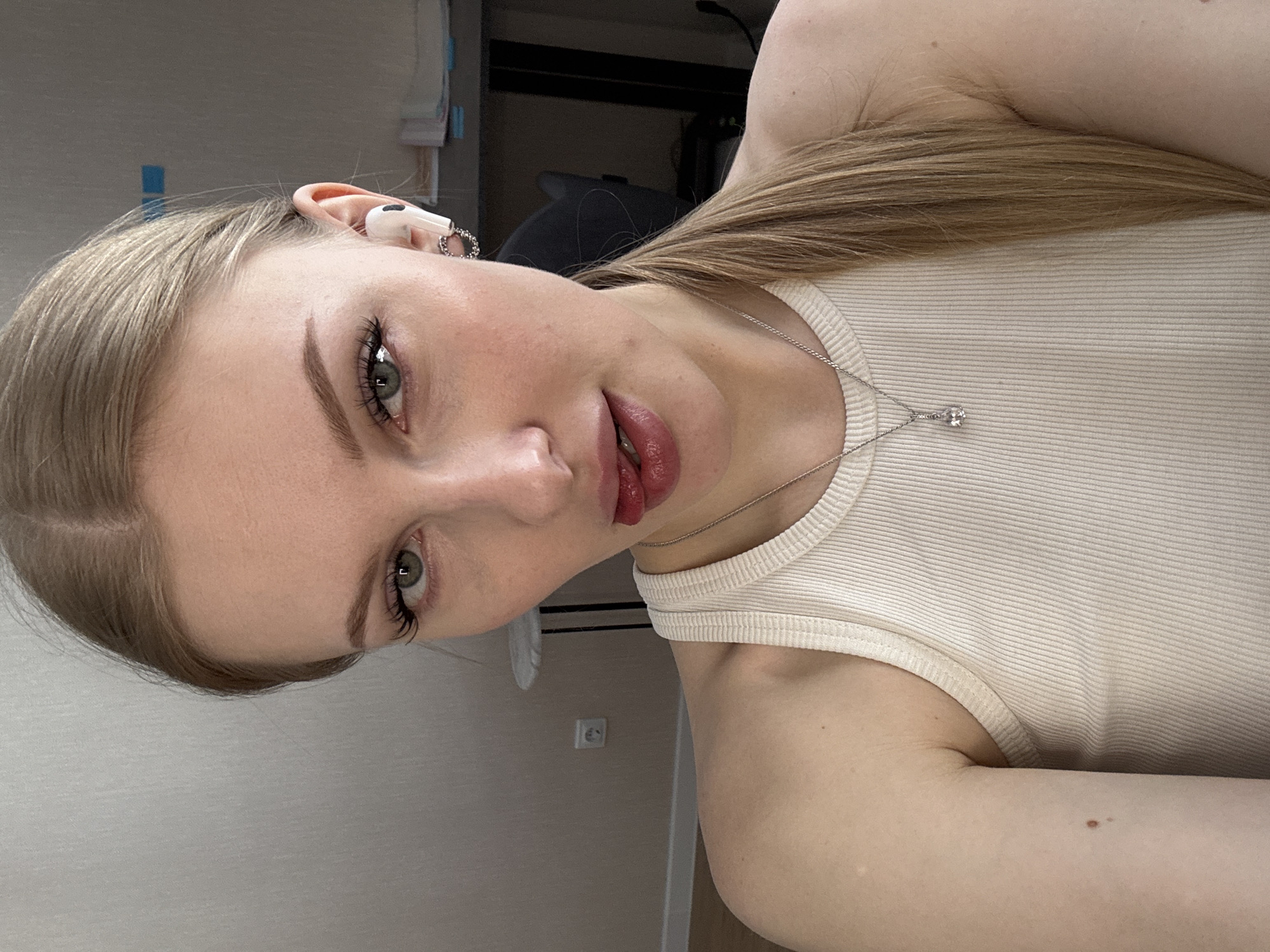 A young woman with light blonde hair, wearing a white tank top and a silver necklace, is posing for a selfie. Her head is tilted to the left. She has light skin, green eyes, and is wearing light makeup. She has earphones in her left ear. The background includes a beige wall, a power outlet, and some office furniture. This photo was most likely taken indoors. There is no evidence of this image being located in Perm, Russia.