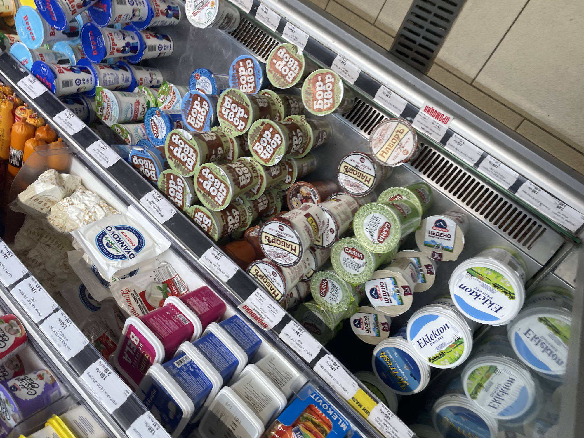 This media file captures a close-up, high-angle view inside a refrigerated display case in a supermarket in Ihtiman, Bulgaria. The scene is dominated by various dairy products, primarily yogurts, cheeses, and dairy spreads, neatly arranged on tilted shelves for customer visibility.
The shelves are packed with numerous brands and types of products. In the upper left, there are stacks of "Верея" (Vereya) brand cups with blue lids. Below them and across the middle, many small tubs are visible, including "Бор-Чвор" (Bor-Chvor), "Наборъ" (Nabor), "Саяна Цедено Мляко" (Sayana Strained Milk), and "Олимпус Цедено Мляко" (Olympus Strained Milk). On the right side, there are several "Еклектон Delicacy" (Eklekton Delicacy) tubs with blue and green designs, and a "Yo Fresh" product. Towards the bottom left, various packaged cheeses are seen, such as "Дианково" (Dyankovo) in white packaging and other items in pink ("Бела" - Bela) and blue ("Акади" - Akadi) rectangular containers. A "Млековит" (Mlekovit) product is also visible at the bottom.
Price tags are prominently displayed along the front edge of the shelves, showing prices in Bulgarian Lev (лв.). Some tags also feature product descriptions like "КРЕМА СИРЕНЕ ОЛИМПУС" (Olympus Cream Cheese), "ОГРАНЕ КОТТЕДЖ" (Cottage Cheese), "МАРГАРИН ВЕСЕЛ" (Vesel Margarine), and "КИСЕЛО МЛЯКО" (Sour Milk). A red label with "ПРОМОЦИЯ" (Promotion) indicates a sale item. No people or specific activities are visible, suggesting a snapshot of merchandise display. The indoor setting and commercial refrigeration unit indicate a grocery retail environment.