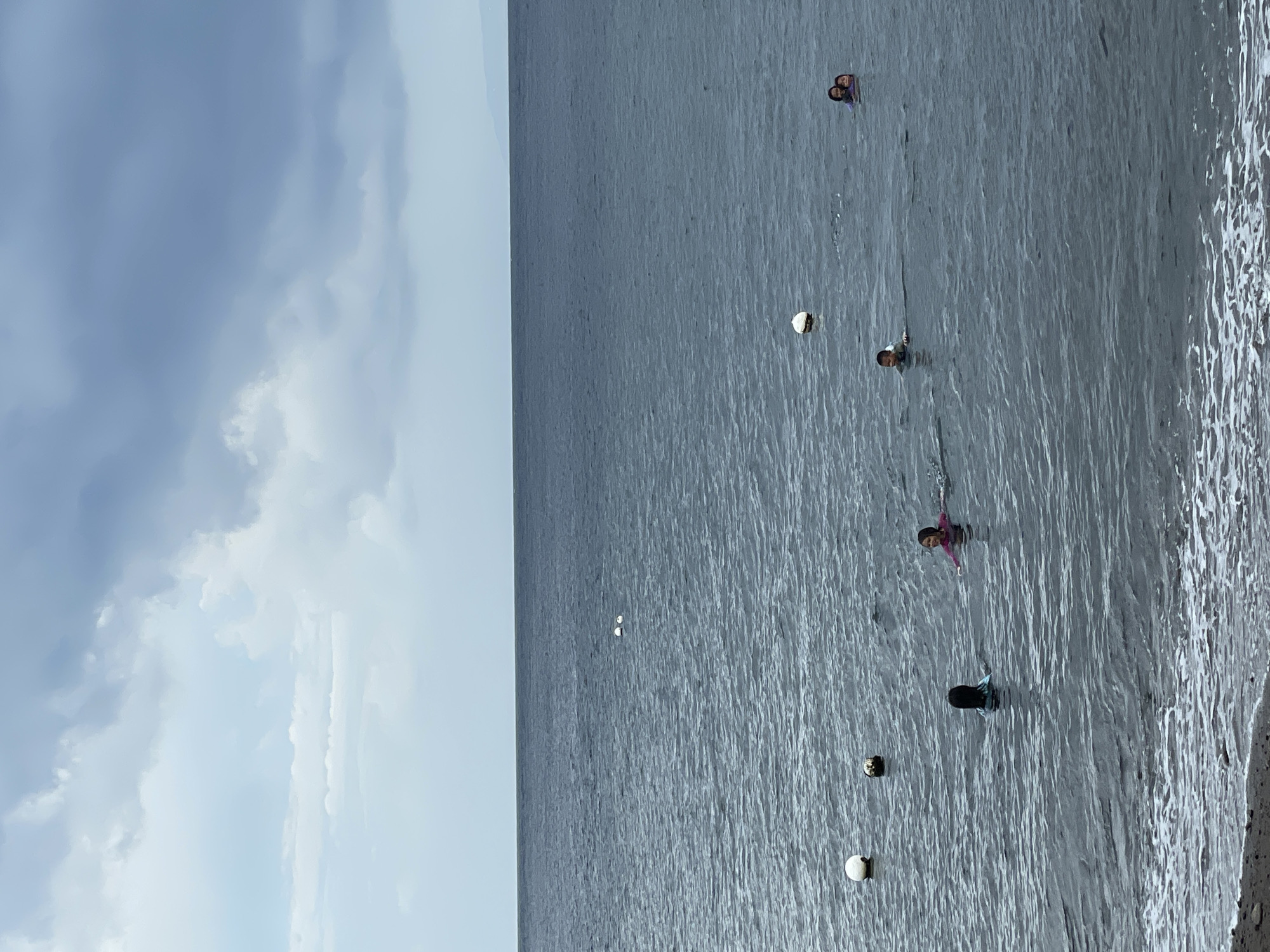 The image shows a body of water with several people swimming and some buoys floating. The sky is overcast with dark clouds. The water is dark blue and appears to be choppy. In the foreground, there is a small strip of beach with some rocks and pebbles. In the middle ground, there are five people in the water. One person in the far left of the frame is wearing a blue top. To their right, two people are swimming together, one wearing a pink top and the other a purple top. Further to the right, another person is swimming, wearing a blue top. In the far right of the frame, two people are swimming together, one wearing a purple top. There are at least three buoys visible in the water. One white buoy is in the lower left corner of the frame. Another white buoy is to the right of it, and a third white buoy is further out in the water. Two smaller white objects, possibly buoys or birds, are visible in the upper middle section of the frame. The horizon line is visible in the distance, with a faint landmass visible on the far right. The image appears to have been taken from the shore. This scene is consistent with the coastal areas of Dauin, Philippines, which is known for its beaches and water activities.