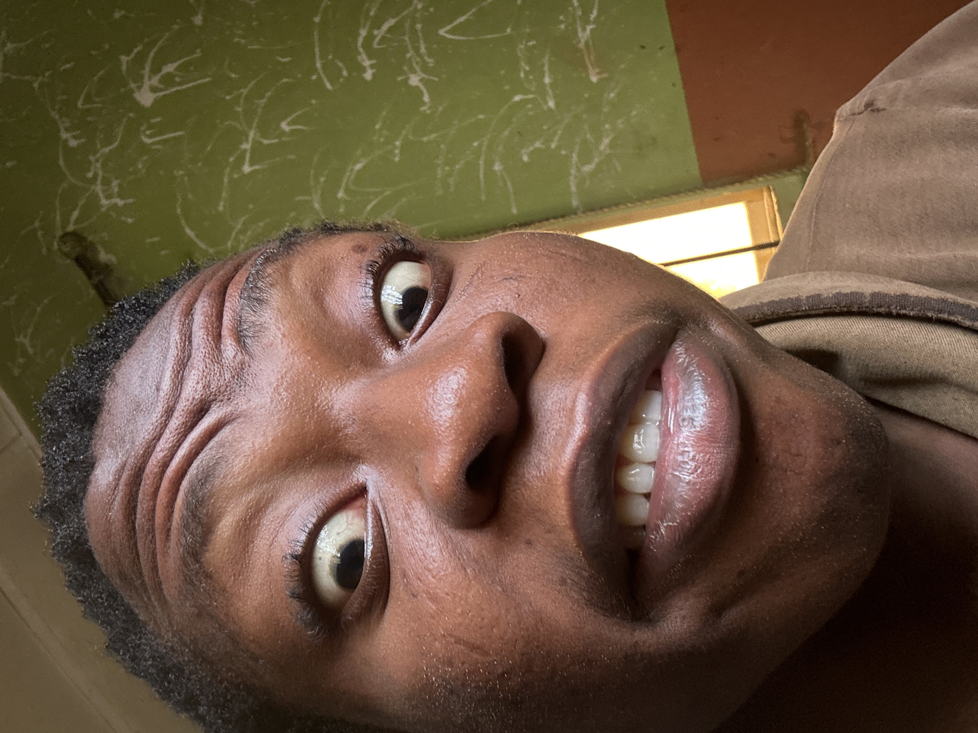 A close-up, downward-angled selfie of a Black person. The subject is positioned facing the camera with a slightly upward gaze. The face features a dark complexion, visible wrinkles on the forehead, and a partial view of white teeth. The background shows a green wall with white markings and a glimpse of a window frame. The location is in the city of Unknown, Unknown.