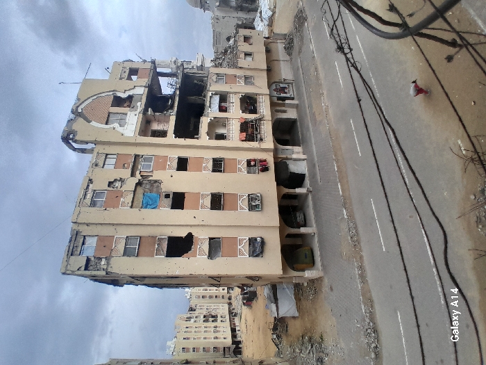 The image depicts a severely damaged urban street scene in Khan Yunis, Palestinian Territory, under an overcast sky. A prominent multi-story residential building, approximately seven floors tall, occupies the center, exhibiting extensive structural breaches, particularly on its upper right side, and numerous impact marks or bullet holes across its light-colored facade. Many windows are shattered or missing; some show hanging clothes. The ground floor features arched entryways. To the left, another similar, less damaged apartment block stands amidst debris and corrugated metal shelters. To the right, a vast area of rubble indicates significant destruction, with the dome of a building partially visible beyond. In the foreground, an asphalt road with faded white markings is traversed by dark utility cables. A single individual, dressed in light pants and a dark top, is observed bent over on the street, near the right foreground.