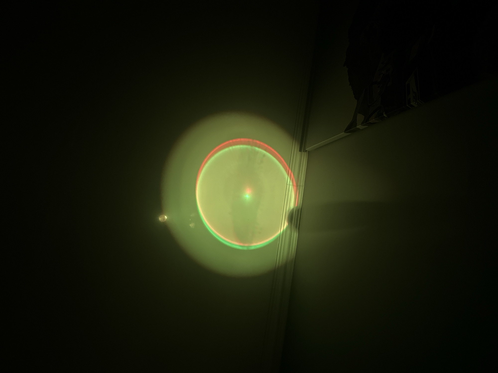 The scene depicts an interior space, likely a room, in the city of Unknown, Unknown. The environment is predominantly dark, indicating low ambient light conditions. A prominent, large, somewhat elliptical light pattern is projected onto the light-colored ceiling and the upper portion of an adjacent wall. This projection features a central bright point of light, surrounded by a diffuse green glow. Within this green area, two distinct, slightly offset concentric rings are visible: an inner green ring and an outer red ring. The outer perimeter of the primary illuminated area also exhibits chromatic fringing, with a green tint on the left side and a red tint on the right.
Below the illuminated ceiling, a white or light-colored architectural molding (e.g., crown molding) runs horizontally along the wall-ceiling junction, partially intersected by the light projection. Further down the wall, a dark, elongated object resembling a curtain rod or support is visible on the right side, extending vertically, with dark fabric or material draped over its lower section, descending into the lower right corner of the frame. A faint shadow, possibly from the projection source or an obstruction, is cast onto the wall below the main light pattern, slightly left of center. No individuals or discernible actions are present in the image.