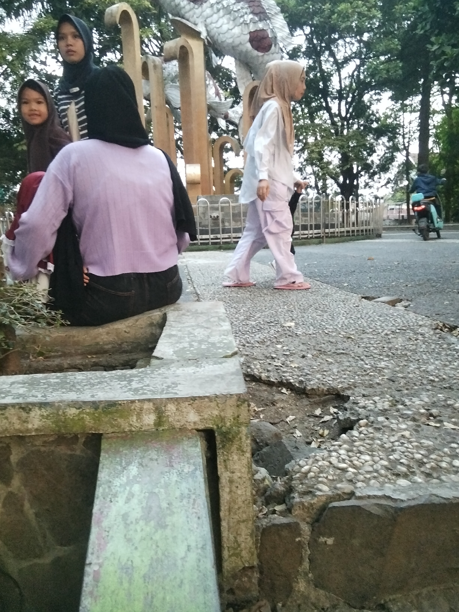 The image depicts an outdoor scene in the city of Cisaat, Indonesia. In the foreground, a person wearing a purple sweater and dark pants is seated on a stone structure. To their left, another person in a dark hijab is visible. To the right of the seated person, a woman in a white shirt and light pink pants, also wearing a light brown hijab, is walking. In the background, trees line a street where a person on a turquoise and black motorcycle is riding away from the camera. A decorative sculpture and a metal fence are also present in the background. The ground is paved with small stones and some patches of cracked concrete.