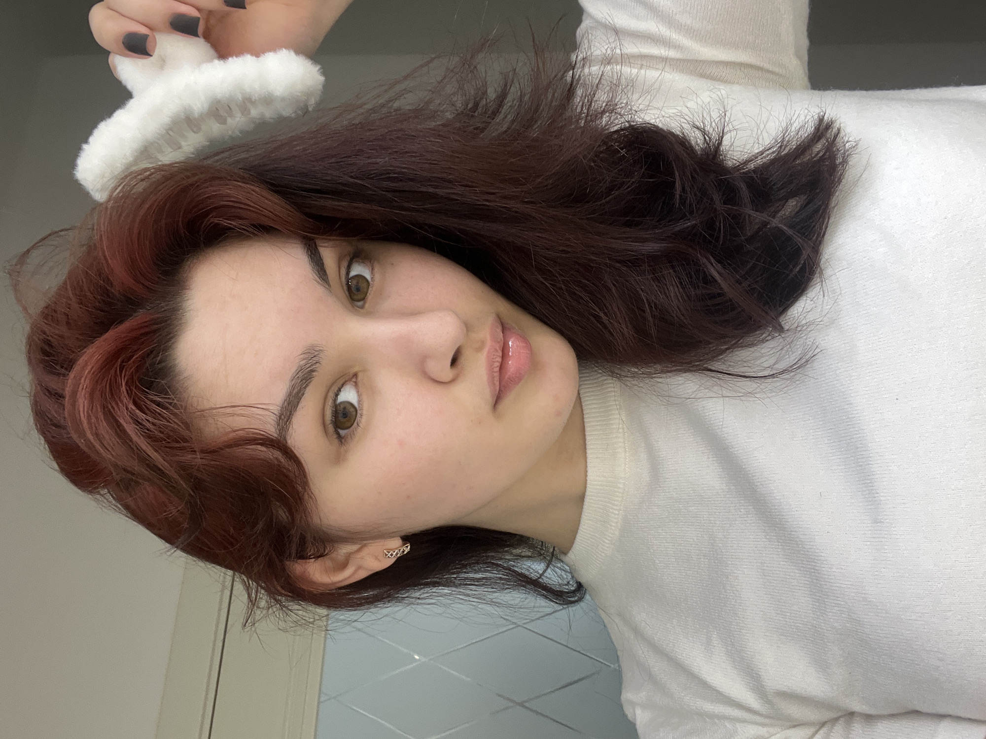 A medium-shot portrait of a woman with auburn-colored hair wearing a white sweater, with a white fuzzy hair clip. Her hand is visible above her head, with dark painted nails. Her eyes are hazel in color. She has a gold earring in her left ear. The background consists of a door and a blue-and-white patterned wall. There is no indication of the location being Neftekamsk, Russia.