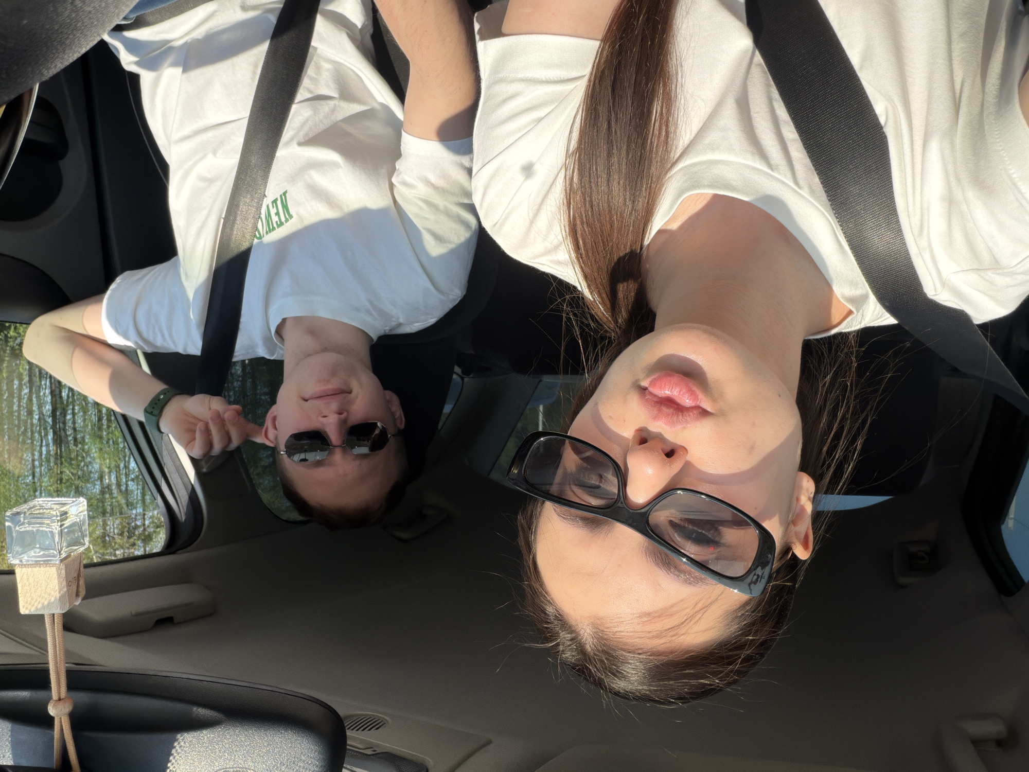 Two people wearing sunglasses and seatbelts are sitting in a car. The woman is wearing black-framed glasses, and the man is wearing metal-framed sunglasses. Both are wearing white t-shirts. The location appears to be Новое Мартьяново, Russia, judging from the environment visible through the car window. An air freshener hangs from the rearview mirror.