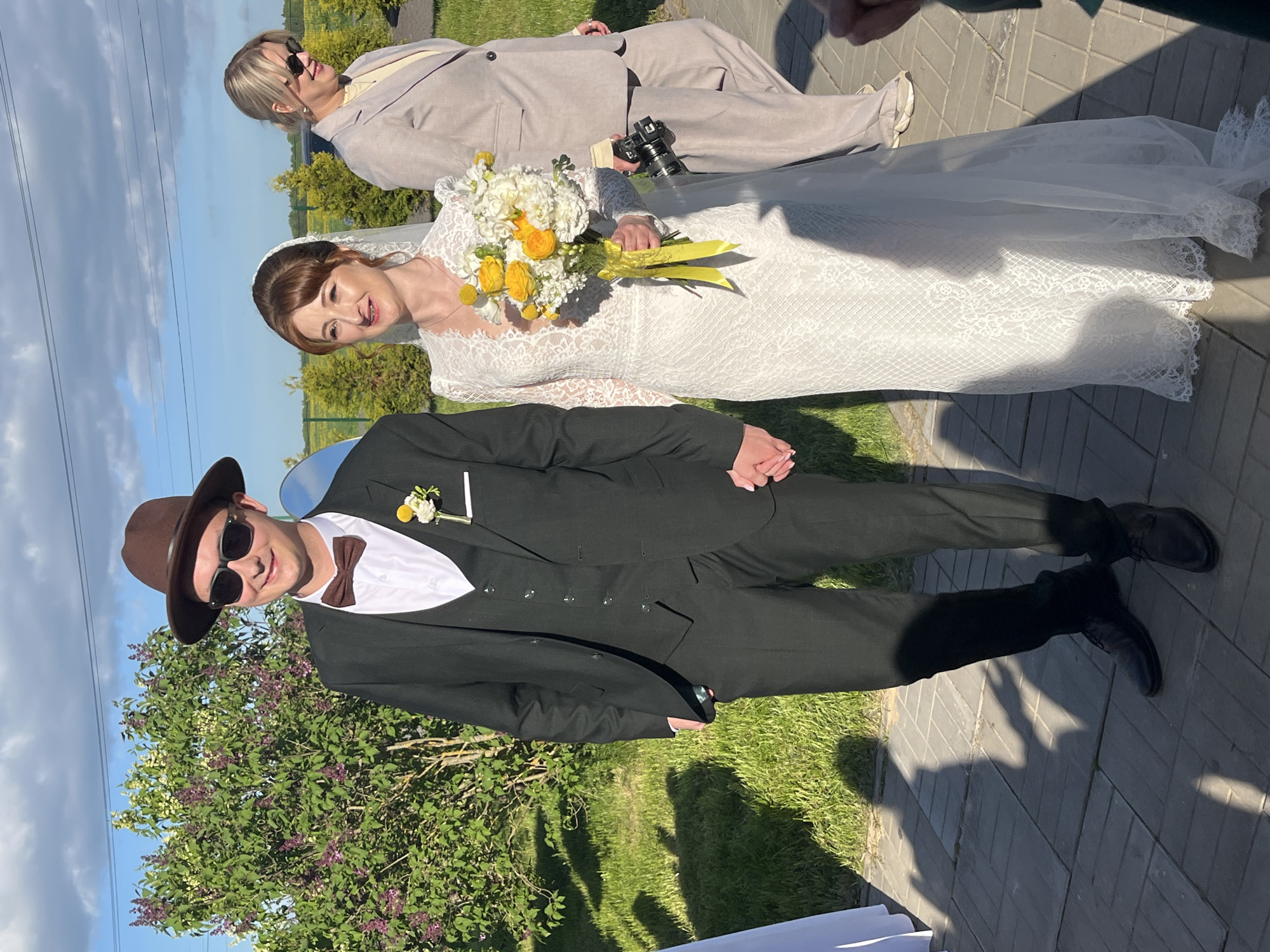The image shows a bride in a lace wedding dress holding a bouquet of yellow and white flowers, standing next to a man in a black suit, brown bowtie, and brown fedora. A person in a light grey suit is also present, lying on the ground and holding a camera. The location appears to be outdoors, with greenery including trees and grass in the background and a paved area. Based on the image, it is not possible to verify the location in the city of Alopovo, Russia.