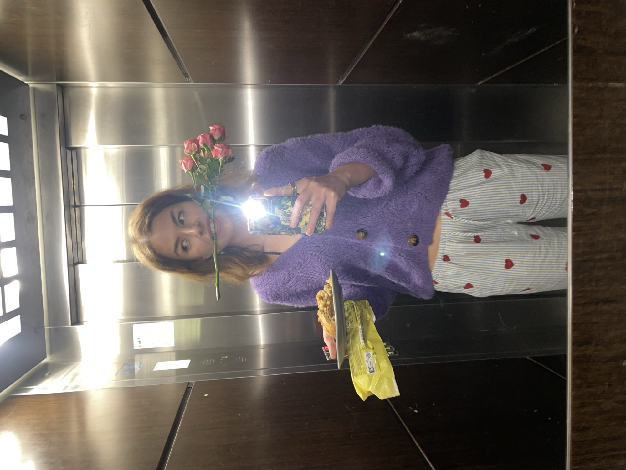 A person, likely female, with light brown hair, is captured in a mirror selfie taken inside an elevator. She is wearing a fuzzy purple cardigan and light blue and white striped pajama pants with red heart patterns.
In one hand, she holds a small bouquet of pink roses, and in the other, she is taking the photo with a smartphone that has a decorative case, its flash brightly illuminating the scene. She also appears to be holding a pastry or muffin in yellow packaging.
The setting is an elevator with reflective metallic walls, which appear to be stainless steel, mixed with dark wooden panels. Elevator control panels with various buttons and indicators are visible on the left side of the reflection. The image is taken from a low angle relative to the subject's face, making the ceiling of the elevator also visible. The activity is a casual mirror selfie, possibly documenting an everyday moment.
No specific time of day is discernible, but the bright camera flash is a prominent feature. No clearly legible text is visible on the elevator controls or the food packaging. The scene takes place in Boreti, Montenegro.