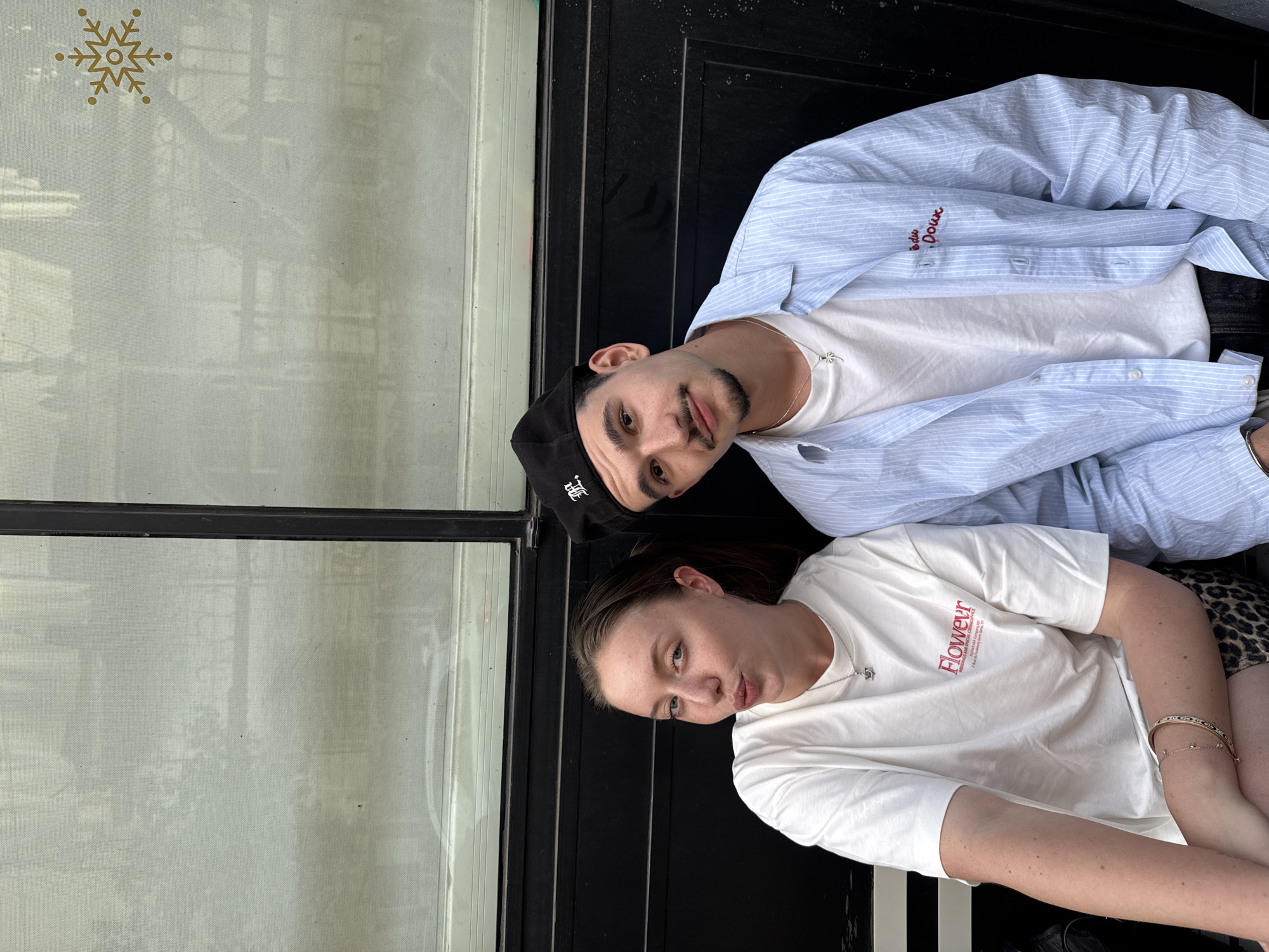 Two people are posing in front of a building in what appears to be the city of Tel-Aviv, Israel. The building's window has a snowflake decal on it. A man is on top, wearing a white t-shirt, a partially unbuttoned light blue striped shirt, and a black baseball cap. He has a short beard and mustache. A woman is below him, wearing a white t-shirt with the word "Flower" on it. She has a necklace, and on her wrist, she wears a beaded bracelet and an additional bracelet. They are leaning against a black vertical structure.