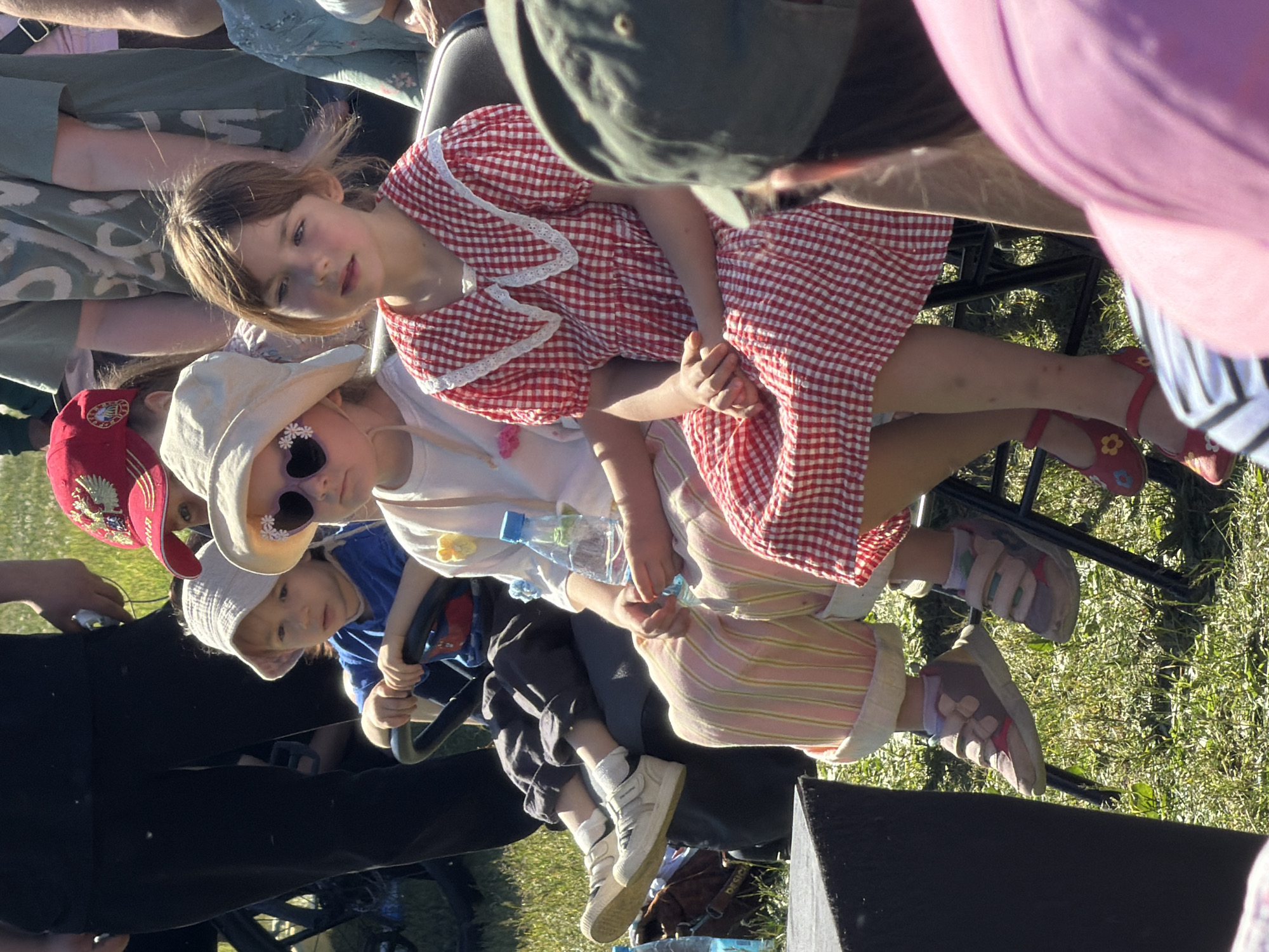 A detailed view in Khimki, Russia, captures three young children seated in the foreground of an outdoor, sunlit environment with grassy ground.
From left to right:
1. A young child, appearing male, wears a light beige bucket hat, a blue t-shirt with a graphic, dark grey pants, and white sneakers with white socks. The child is seated in a dark-colored, possibly ride-on toy, with a black steering wheel visible in their hands. Their gaze is directed towards the left. A clear plastic water bottle is partially visible next to their right leg.
2. A young girl, positioned in the center, wears a wide-brimmed light beige sun hat, dark heart-shaped sunglasses with white floral embellishments, a white t-shirt adorned with a small yellow flower, and pink and yellow vertically striped pants. She is holding a blue plastic water bottle in her lap. Her feet are encased in multi-colored (pink, grey, white, blue) sneakers secured with velcro straps, dangling above the ground. She looks forward.
3. A young girl, positioned on the right, wears a red and white gingham-checked dress featuring a white lace Peter Pan collar and short puffed sleeves. She has dark hair and her hands are clasped in her lap. Her feet wear red Mary Jane-style shoes with yellow floral decorations, also dangling above the ground. Her gaze is directed towards the left foreground.
The children are seated on individual dark-colored, lightweight chairs. In the immediate left foreground, a black rectangular object, possibly a speaker or stage element, is partially visible.
Behind the leftmost child, the lower torso and legs of an adult wearing dark pants are visible. This adult holds a white object, possibly a smartphone, in their right hand. Further into the mid-ground and background, several other individuals are present, mostly out of focus. Visible elements include a person wearing a red cap with an emblem, another wearing a grey-green patterned t-shirt, and various partial views of other attendees, suggesting a public gathering or event. In the far right foreground, the back of a head wearing a green baseball cap and part of a shoulder in a pink top are visible. The scene is illuminated by direct sunlight, casting shadows on the ground.