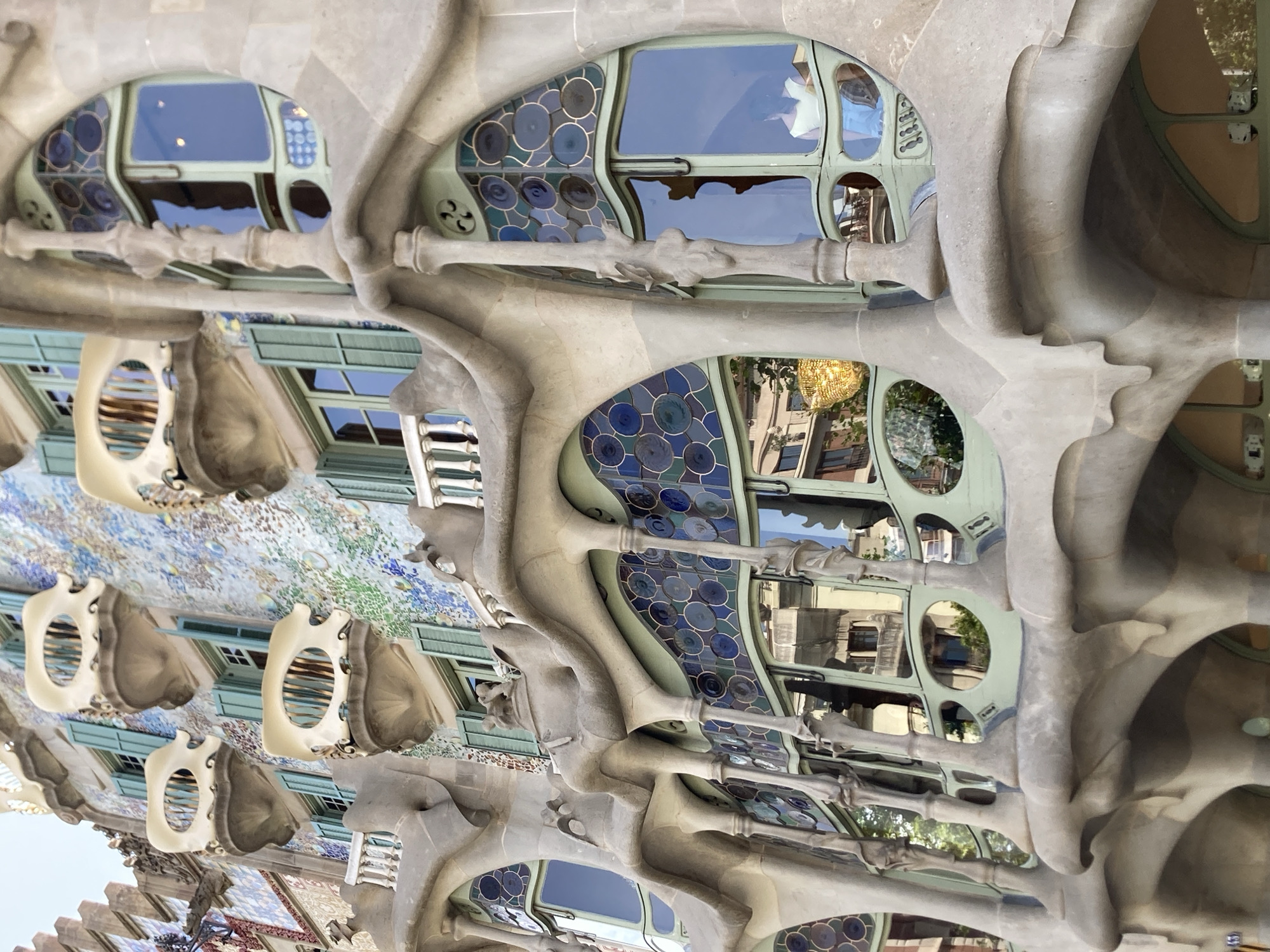 This image showcases the facade of Casa Batlló in Barcelona, Spain, a modernist masterpiece by Antoni Gaudí. The perspective is looking upwards at the building's iconic organic-inspired architecture. The facade features a wave-like stone structure with various window openings. Some windows are rounded and adorned with stained glass mosaics in shades of blue and green, resembling water bubbles or natural forms. Other windows are larger, rectangular panes set within intricate frames.
Suspended from the building's exterior are several white, bone-like structures that appear to be balconies or decorative elements. These elements are hollow and have a sculptural quality. The stonework itself is fluid and curved, mimicking natural shapes like bones or flowing drapery. Reflections are visible in the windows, suggesting a bright, possibly sunny day. The overall impression is one of artistic innovation and a harmonious blend of architecture and nature. There is no visible text in the image.