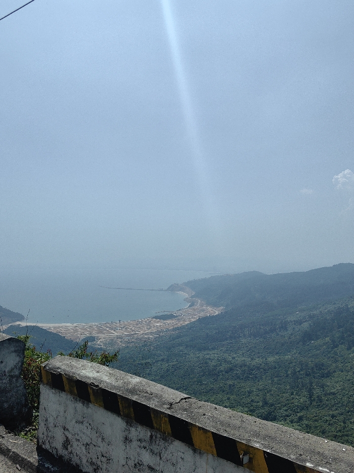 The image displays an expansive high-angle view of a coastal landscape within Hải Vân Ward, Vietnam, under a bright, hazy sky. In the immediate foreground, a weathered concrete road barrier or retaining wall is visible, featuring a white base with diagonal black and yellow stripes. Beyond the barrier, a dense green forested mountainside slopes down towards a large bay.
The bay's waters appear a pale blue-grey, extending towards the horizon where they meet a distant, hazy coastline. Along the curving shoreline of the bay, an extensive area of land appears developed or under development, characterized by numerous light-colored, rectangular plots or structures. A prominent, dark-colored pier or breakwater extends into the bay from the right side of the frame, partially enclosing an area of water. Several small, dark objects consistent with fishing boats are faintly visible in the water near the shore.
The background consists of multiple layers of lush, verdant green mountains and hills receding into a atmospheric haze. The sky is a light, clear blue, with a distinct, bright sun flare traversing diagonally from the upper left towards the center-right. A small, white cumulus cloud is present in the upper right quadrant. No people, specific actions, or interactions are discernible.