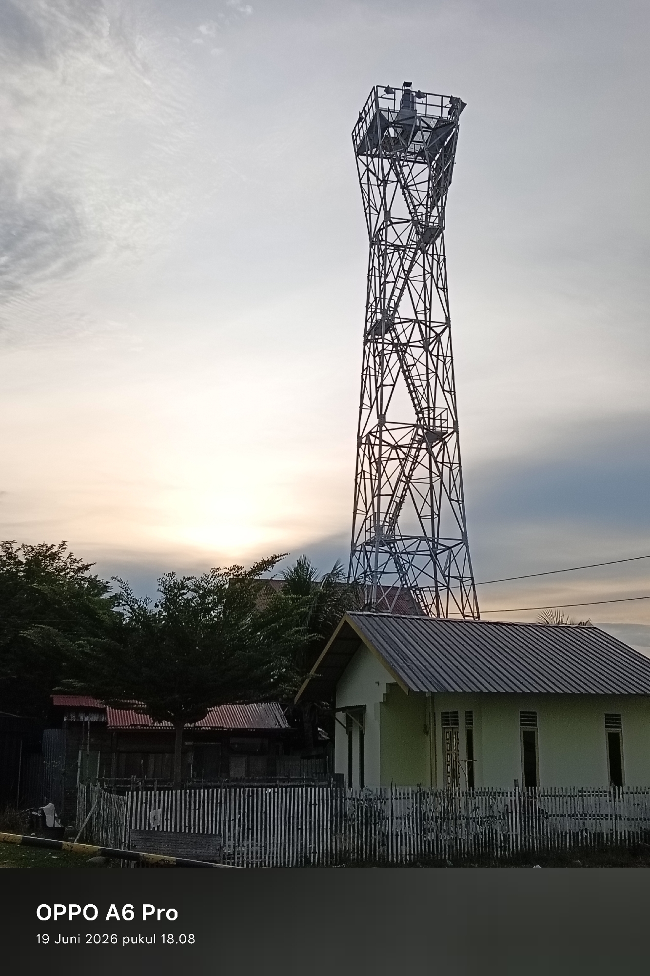 This is a photograph taken in Lhokseumawe, Indonesia, at 18:08 on June 19, 2026, using an OPPO A6 Pro. The scene features a tall, lattice-style telecommunications tower dominating the right side of the frame. In the foreground, a small, light green building with a corrugated metal roof is visible, partially obscured by a white picket fence made of vertical slats. To the left of the building, there are several smaller structures with red corrugated metal roofs, and dense green foliage. The background consists of a hazy sky with the sun setting or rising, casting a warm glow. The image appears to be captured from ground level.