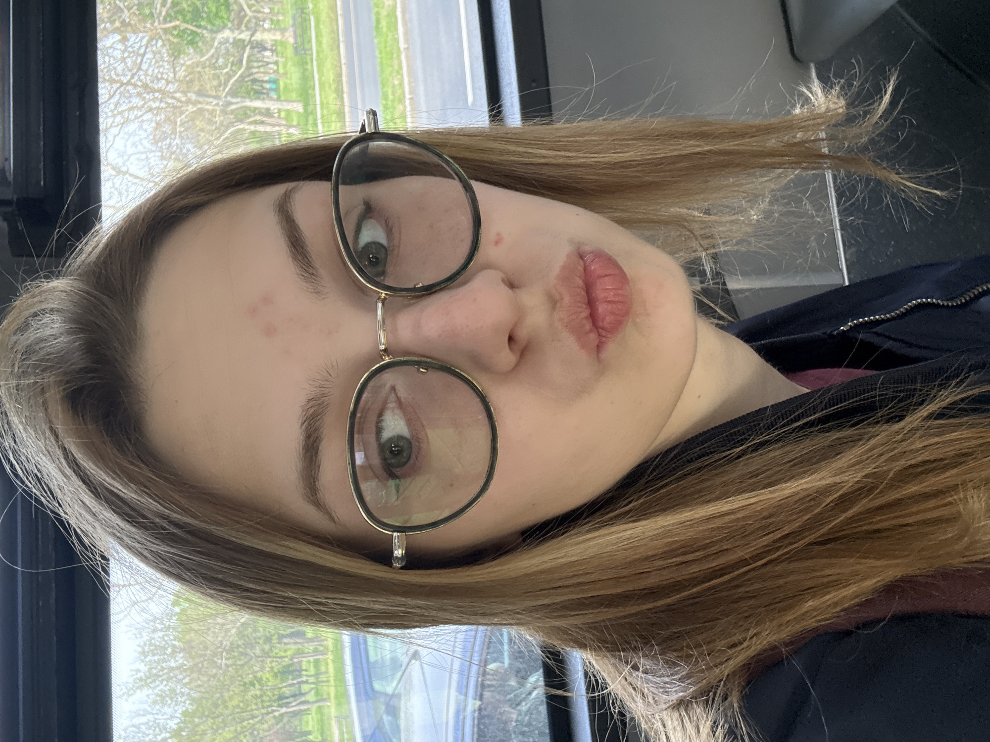 A young woman with light brown hair is wearing glasses and has light skin. She appears to be in a vehicle, possibly a bus, based on the window and the presence of a black frame around it. Outside the window, trees and a road with a vehicle are visible. There is no information in the image that indicates this is located in Belgrade, Serbia.