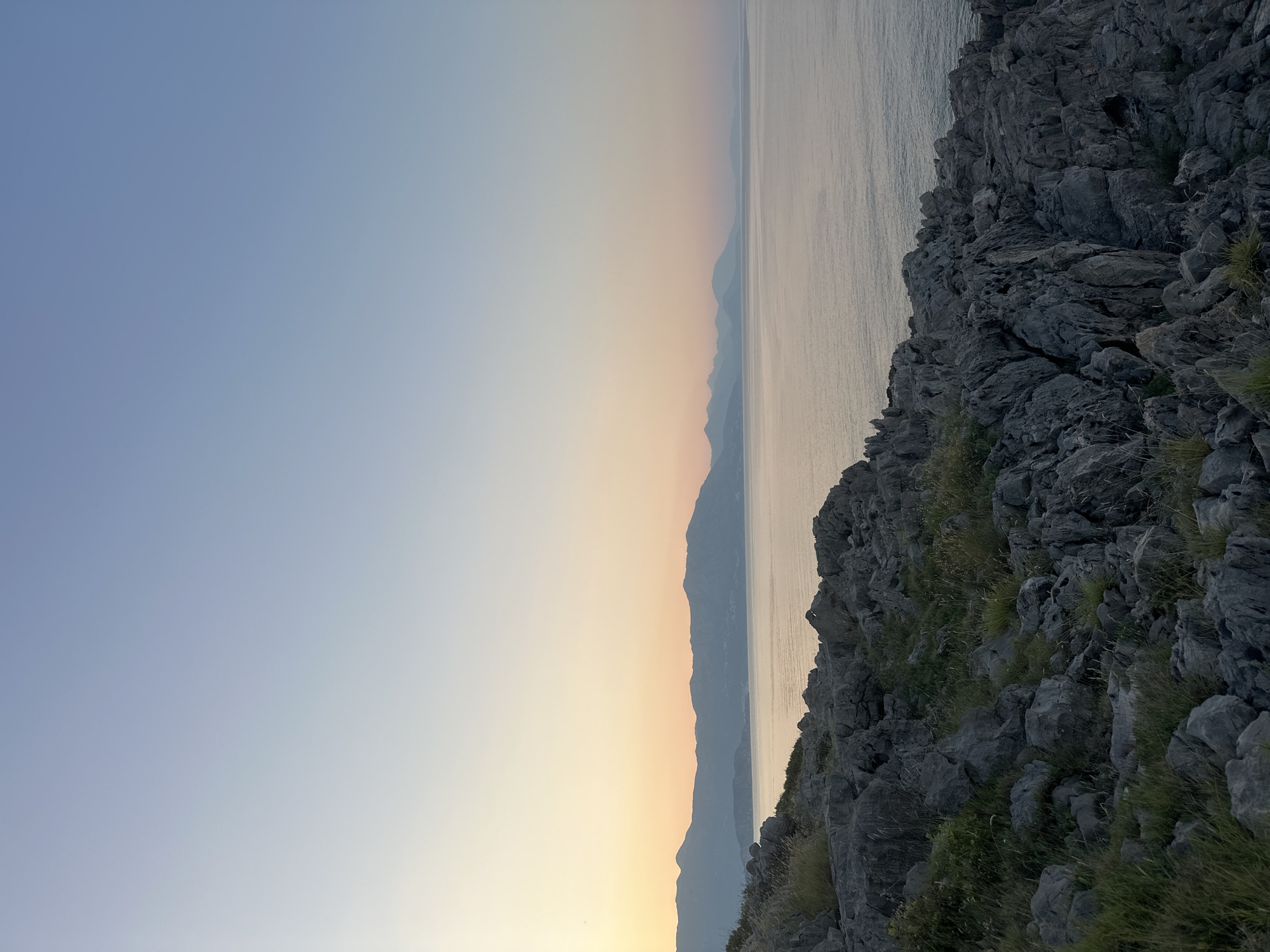 A panoramic view of the sea and coastline at sunset in Dubraljevina, Montenegro. The foreground is dominated by rugged, grey rocks interspersed with dry grass and green vegetation. The sea stretches out to the horizon, with a calm, reflective surface that shimmers under the fading light. In the distance, a chain of silhouetted mountains forms a dramatic backdrop against the pastel hues of the sunset sky. The sky transitions from a soft yellow and orange near the horizon to a pale blue at the zenith. There are no people or man-made structures visible in this natural landscape.