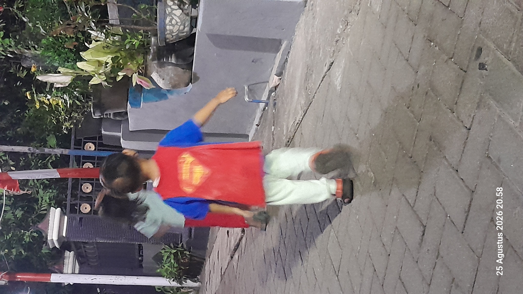 A nighttime, medium shot captures two children walking away from the camera on a paved walkway. The child in the foreground, wearing a blue shirt with a red Superman cape, light-colored pants, and sandals, is positioned to the right of the frame. The second child, to the left and slightly behind, wears a light blue shirt and red pants. Behind the children, a dark fence with circular embellishments runs horizontally. To the right of the fence, several potted plants are visible on a raised concrete structure. The walkway appears to be made of interlocking grey pavers, some of which are cracked. The overall lighting suggests an outdoor environment with artificial illumination. A timestamp in the bottom left corner reads "25 Augustus 2026 20.58," indicating the image was taken on August 25, 2026, at 8:58 PM. The scene is consistent with a residential or public outdoor space in Madiun, Indonesia, given the timestamp and common architectural and environmental features.