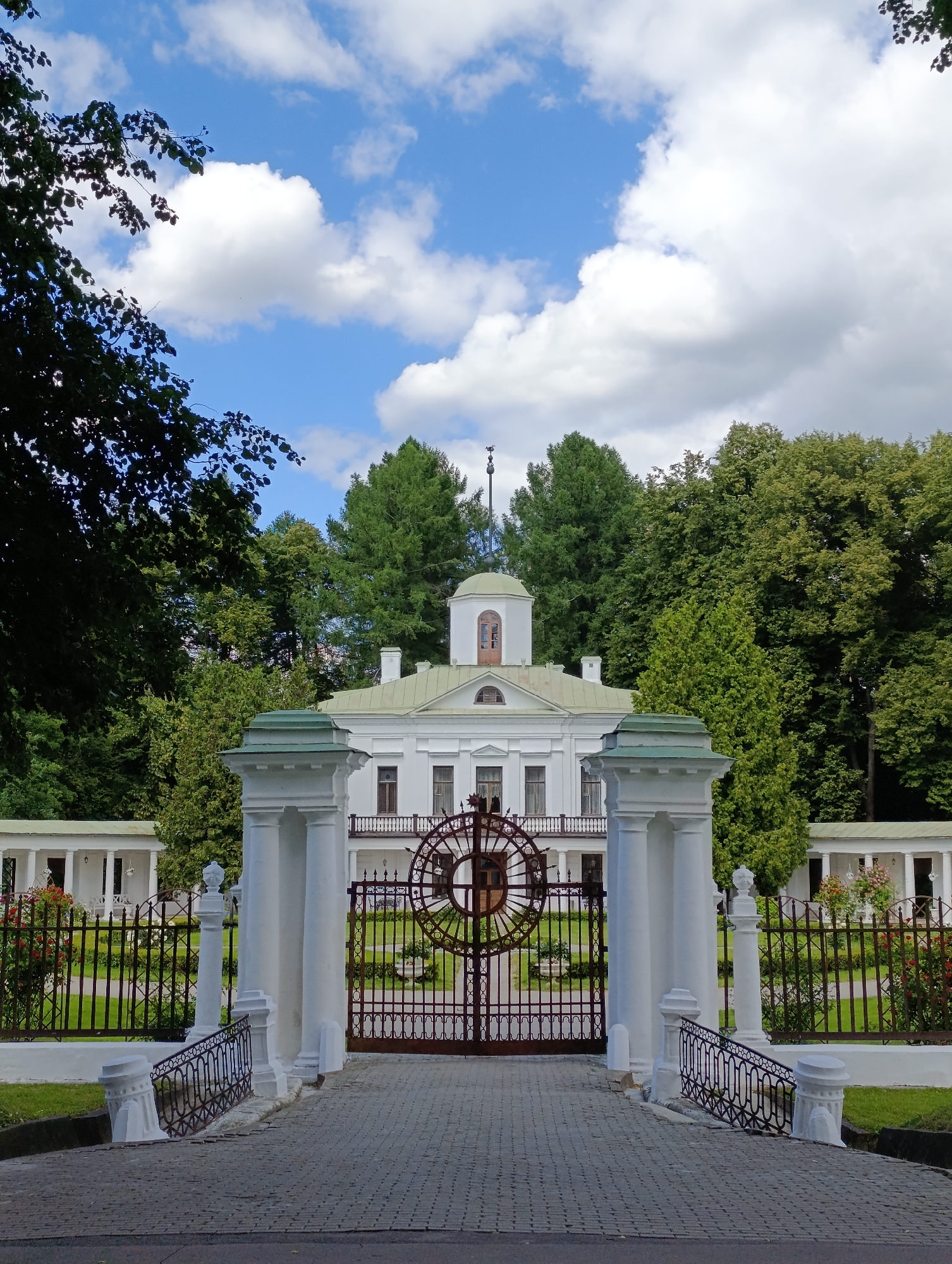 This is a full-shot, vertical, eye-level, outdoor photograph of a grand white estate with a large, ornate gate in the foreground. The estate building is a two-story structure with a green, domed cupola on top, and a balcony with a dark railing. The building has several windows and is surrounded by lush green trees. The gate is made of dark metal with intricate circular patterns and is flanked by large white pillars. A paved walkway leads up to the gate. A black metal fence with decorative elements runs along the sides of the property, with blooming red flowers visible in planters. The sky above is blue with scattered white clouds, suggesting a pleasant daytime with good weather. The scene evokes a sense of tranquility and historical grandeur. The location is identified as Ligachevo, Russia. There is no visible text in the image.