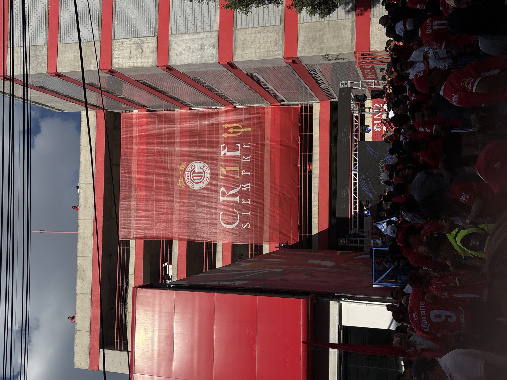 The image shows a crowd of people, many wearing red jerseys, gathered near a red and grey building with a large banner displaying the logo and text "CR13L SIEMPRE" (likely a Toluca FC slogan). The banner covers a section of the building's façade. There's a stage set up with a video screen, suggesting a public event or rally. Power lines are visible in the upper left, and elements of urban infrastructure are present. The location is likely near Estadio Nemesio Díez in Toluca, Mexico.