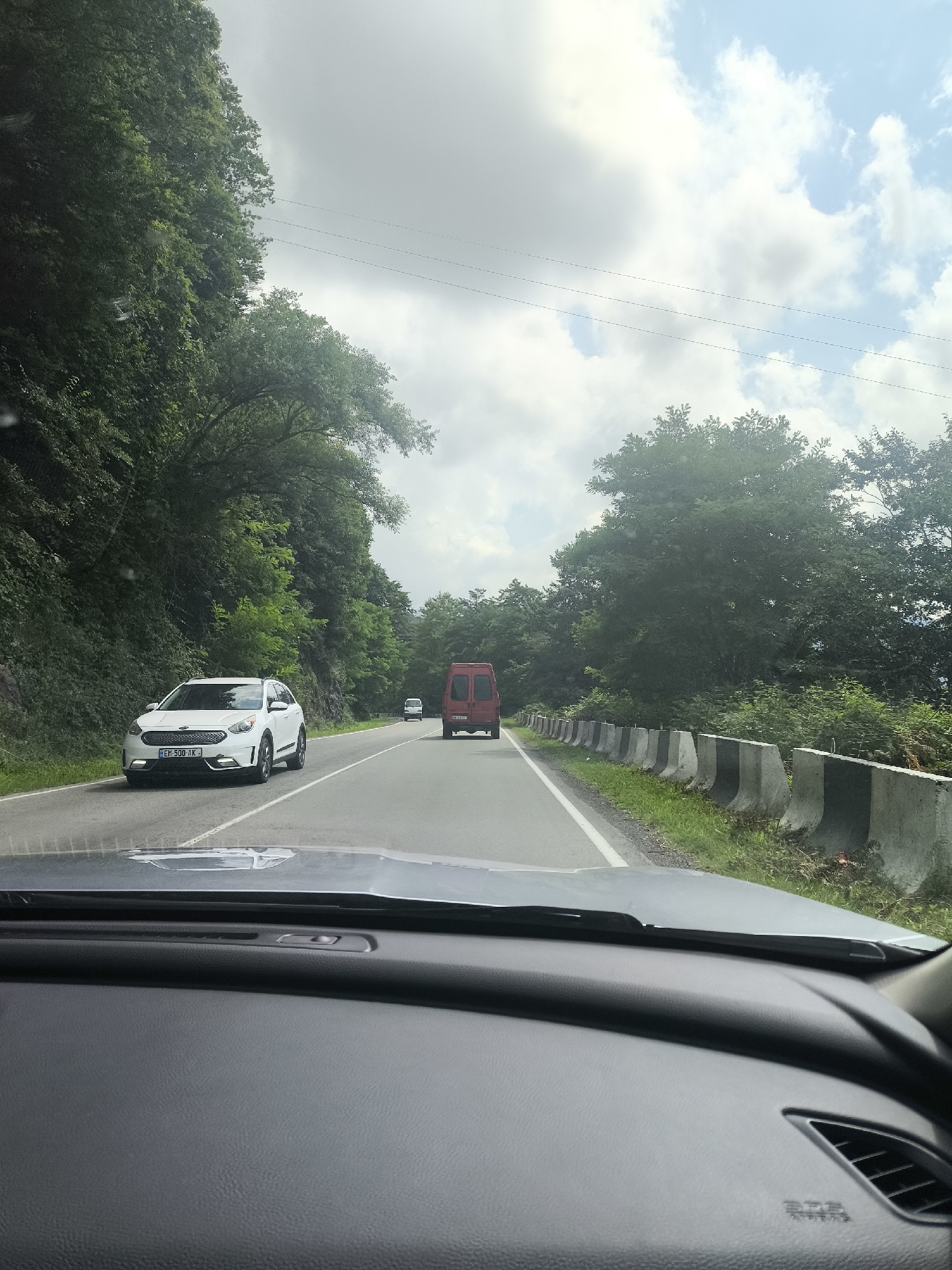 This is a first-person perspective, dashboard-mounted shot of a vehicle traveling on a road flanked by dense green foliage. The road is paved and has a white line marking the lanes. Several vehicles are visible ahead: a white SUV, a red van, and another smaller white vehicle. The SUV is in the left lane, and the red van is in the right lane. The terrain is hilly, with trees covering the slopes on either side of the road. Concrete barriers are present along the right side of the road. The sky is partly cloudy with scattered white clouds against a blue background. The overall impression is of a scenic drive through a wooded, possibly mountainous area. The image was captured on the road to Erge, Georgia.