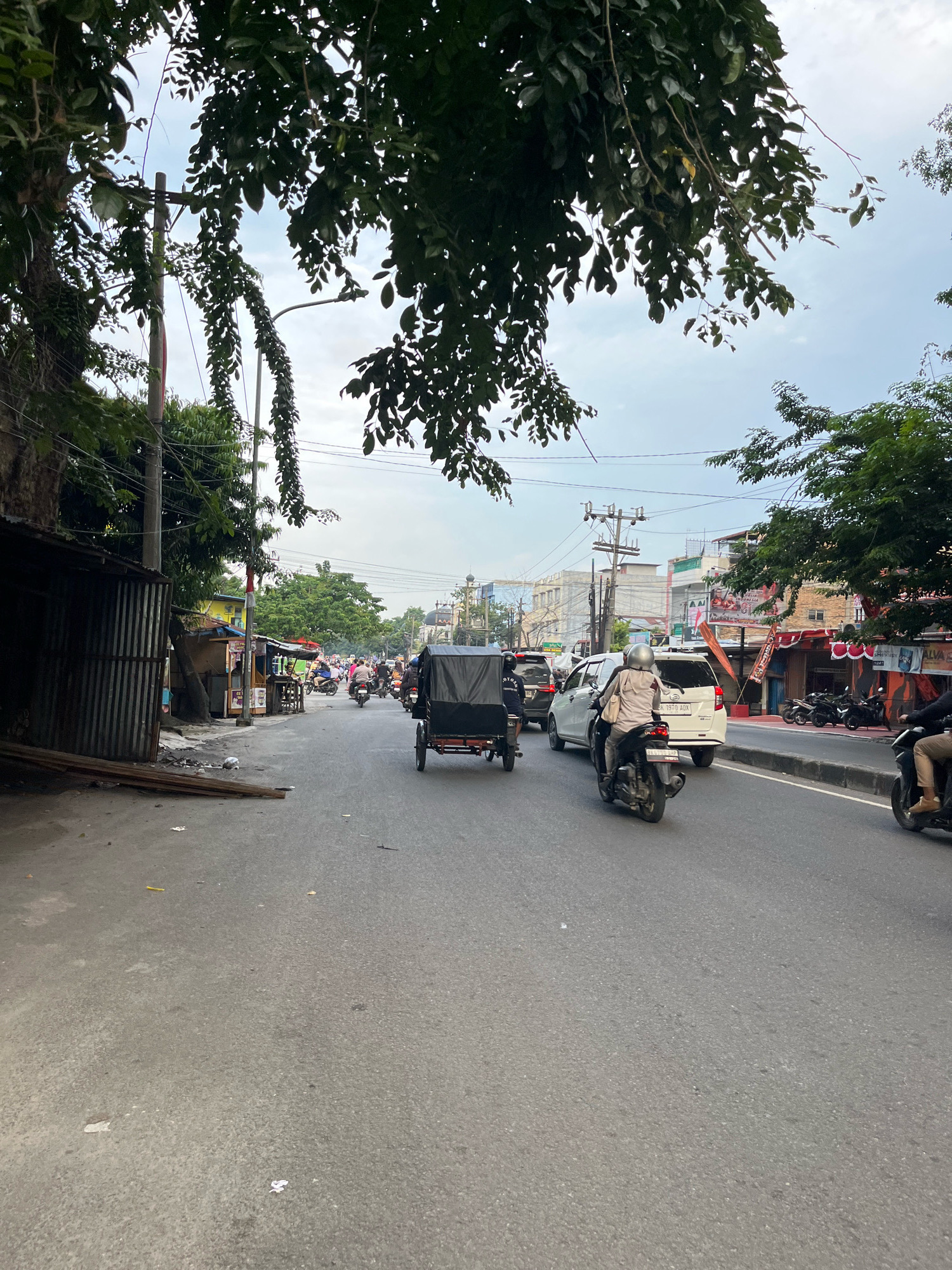 The image depicts a street scene in the City of Medan, Indonesia, with a focus on the road and its immediate surroundings. The asphalt road is busy with vehicular traffic, including motorcycles, a white car, and a trishaw with a black canopy. Several people are visible, mostly riding on motorcycles. On the left side of the road, there is a row of single-story buildings with corrugated metal siding, some appearing to be shops or stalls. Further down the street, larger buildings are visible, including what appears to be a mosque with domes and minarets in the distance. Trees with dense foliage overhang the street from the right and top of the frame. The sky is overcast with light clouds. The overall impression is of an active urban environment with a mix of commercial and residential structures and significant street-level activity.