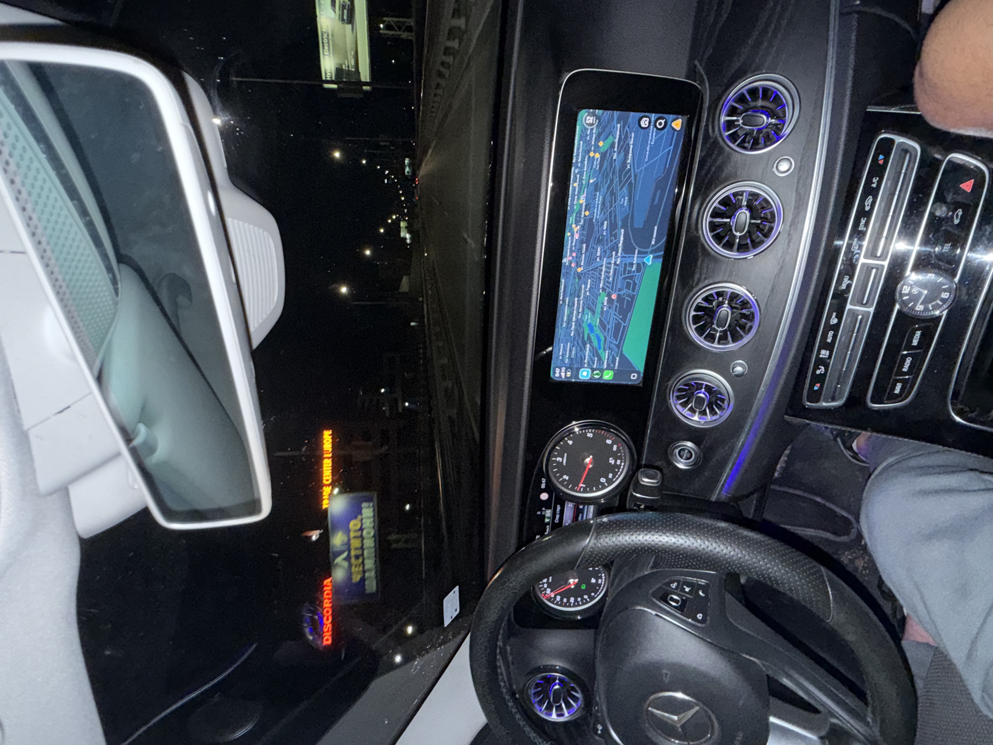 The image depicts the interior of a Mercedes-Benz car, specifically the dashboard and steering wheel area, taken at night. The navigation screen is on, displaying a map of what appears to be an urban area. Through the windshield, streetlights and a sign reading "DISCORDIA TRADE CENTER EUROPE" in Sofia, Bulgaria, are visible. A person's legs are visible in the lower right corner. The car is on the road.
