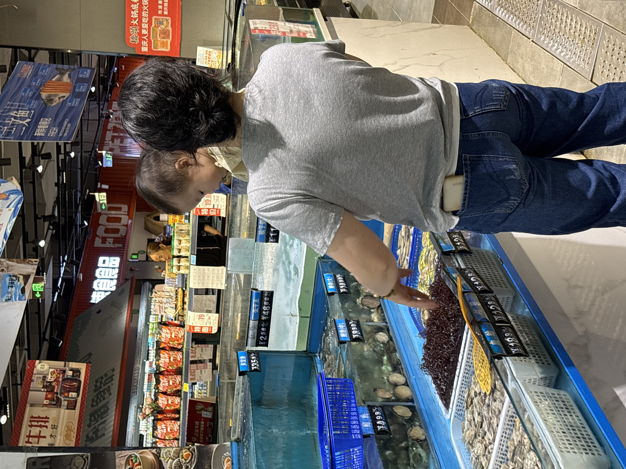 Here is a description of the image:
The photo was likely taken indoors, possibly in a supermarket or grocery store in Jiangjin, China. An adult wearing a light gray t-shirt and jeans is standing near a fish tank or display of seafood. The person appears to be looking at the seafood, perhaps selecting an item to purchase. There are various signs and labels with Chinese characters indicating different types of seafood, with prices listed. There is also a child beside the adult. The location is near other items on the shelves of the supermarket, like hot pot sauces.