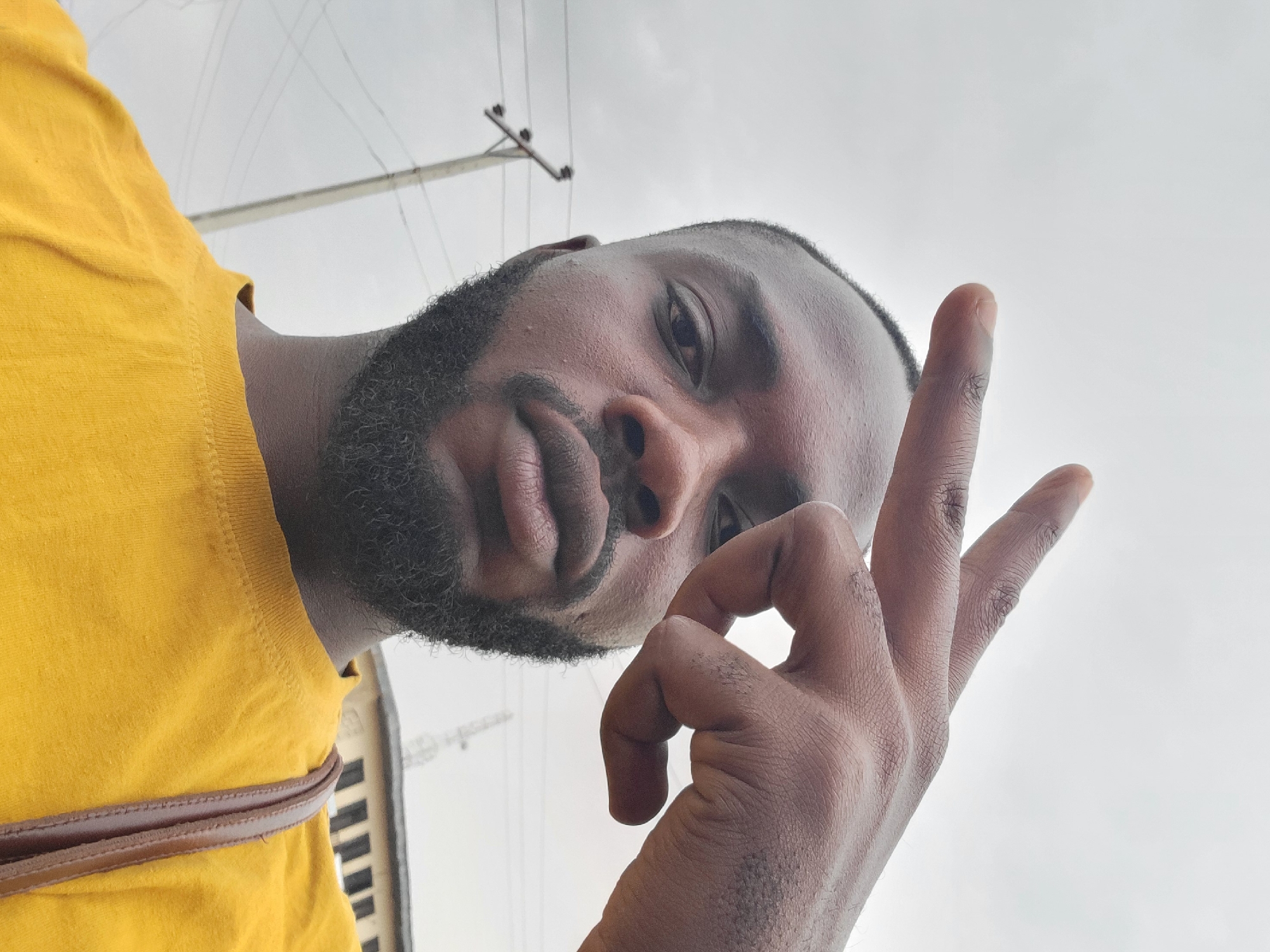 A medium shot of a Black man in Jos, Nigeria, takes a selfie at a low angle. He wears a yellow shirt with a brown strap visible on his left shoulder. The man has dark hair and a full beard. His skin tone is brown. He is making a hand gesture. Behind him, the sky is overcast. An electric pole with wires is visible in the background, as is the top of a building with multiple windows.