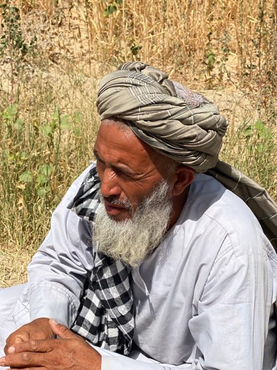 The image depicts an elderly man in Mansurabad, Afghanistan. He is wearing a large, wrapped turban of muted gray and brown tones and a light gray shirt. A black and white checkered scarf is draped around his neck. He has a long, white beard. The background consists of dry grass and vegetation, suggesting an outdoor location. The man is looking slightly to his left with a contemplative expression. His hands are clasped in front of him. The scene is bathed in natural daylight.