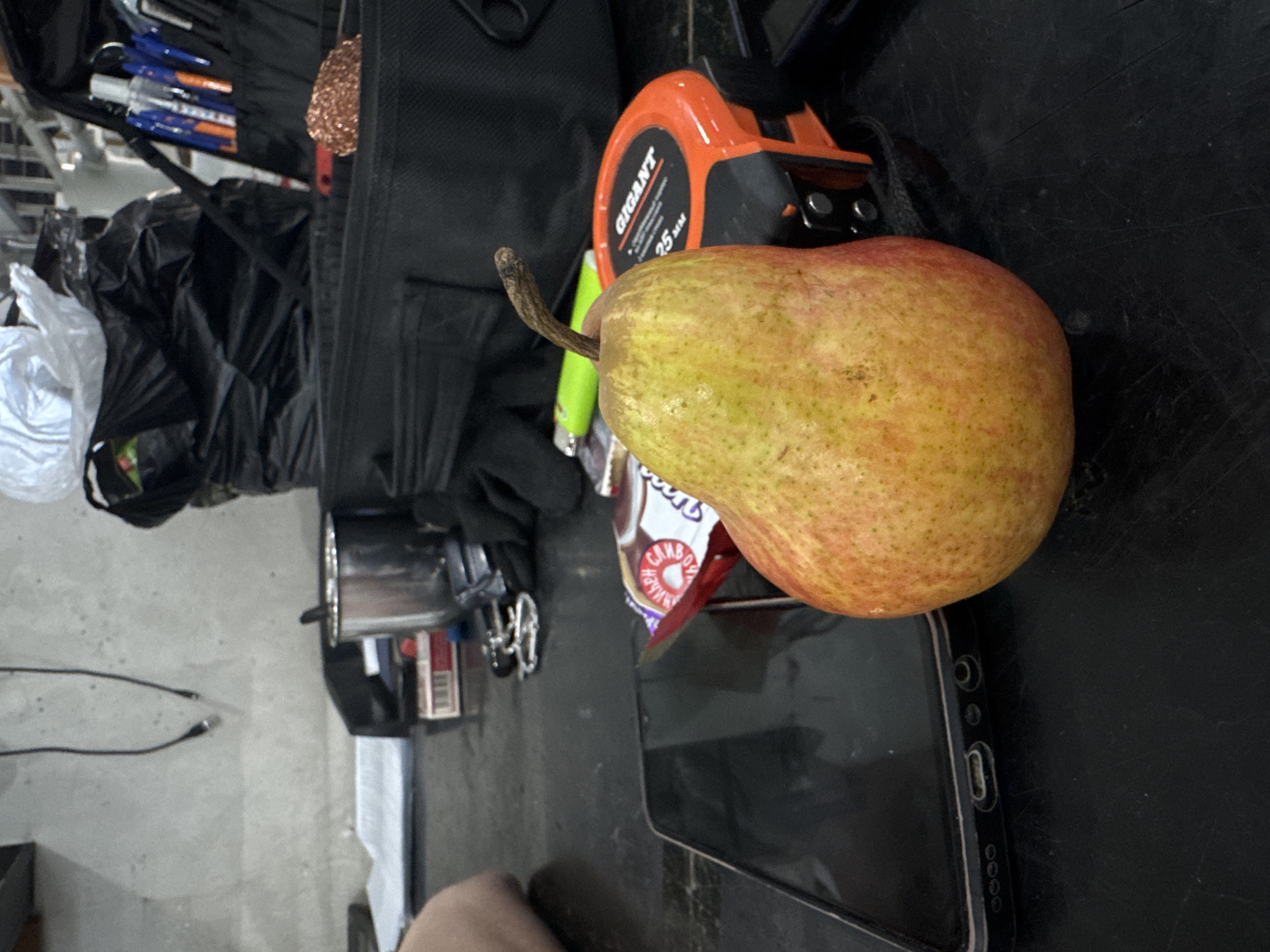 An indoor scene in Dikaya Gar', Russia, displays a collection of utilitarian objects on a dark, worn tabletop or workbench. A yellow-red pear with a visible stem stands upright in the immediate foreground, positioned slightly right of center. A black smartphone is partially obscured beneath the pear, lying flat on the surface. Adjacent to the pear, a red and white snack wrapper with partially visible Cyrillic text, including "ВИКЬЕН С", is present. To the right, an orange and black tape measure, labeled "GIGANT 25 MM", rests beside a neon green lighter. A person's forearm and hand are visible on the far left, resting on the tabletop. In the mid-ground, a silver metal cup, a red and white box resembling a cigarette pack, and a set of keys on a keyring are arranged. A black fabric tool bag, open and containing multiple blue and orange pens, along with a coiled copper object, is positioned on the right. Black gloves are also visible. White papers are stacked on the left side of the table. The background features a light grey concrete wall from which two black cables, terminating in USB-A connectors, hang. Clutter, including black and white plastic bags, is visible in the upper right background, with indistinct metallic shelving beyond.