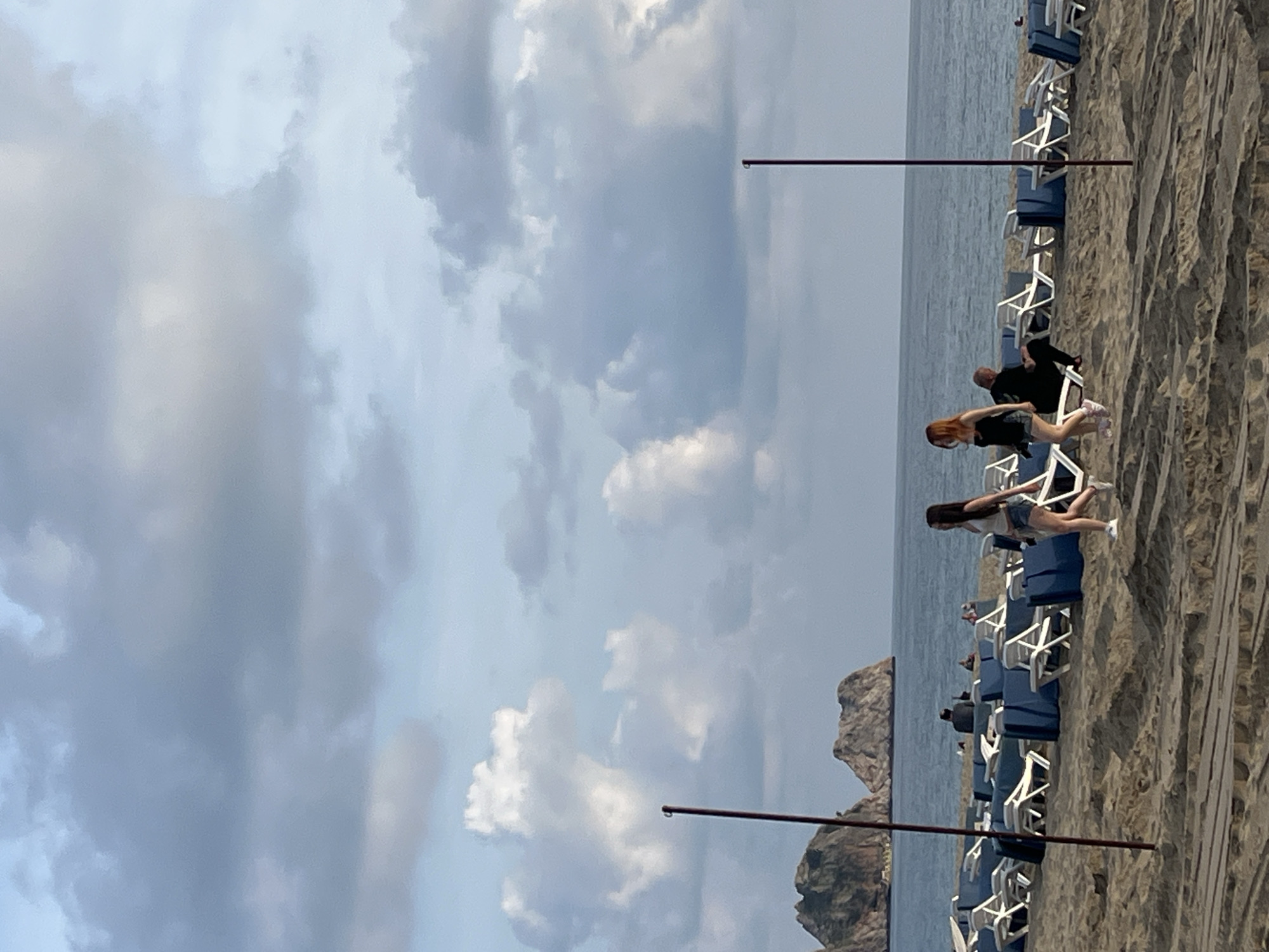 This image, captured in Alanya, Turkey, depicts a serene coastal scene under an overcast sky. The photograph is oriented vertically, showing a wide expanse of sky filled with soft, diffused clouds at the top.
Below the cloudy sky, the calm, grey-blue sea stretches horizontally towards the horizon. Along the coastline, a sandy beach or a wooden boardwalk is visible, where numerous beach chairs are neatly arranged in rows. These chairs feature blue seats and white frames, indicating a resort or public beach setting.
Several individuals are present on the beach. In the foreground, two young women appear to be walking or moving along the sandy path parallel to the sea. One woman has reddish-brown hair and is dressed in a dark top and light shorts, while the other has long dark hair and wears a dark top with light shorts. Further back, another person is seated on one of the beach chairs, and other figures are faintly visible in the distance among the chairs.
Two dark, thin poles or rods extend across the frame, likely part of an overhead structure such as an awning or a railing. One of these poles shows a small water droplet hanging from its underside, suggesting recent precipitation or condensation.
In the background, rising from the sea, a distinctive rocky outcrop or small island adds a natural landmark to the coastal panorama. The soft, even lighting suggests it is daytime, possibly a slightly overcast afternoon. No visible text is present in the image.
