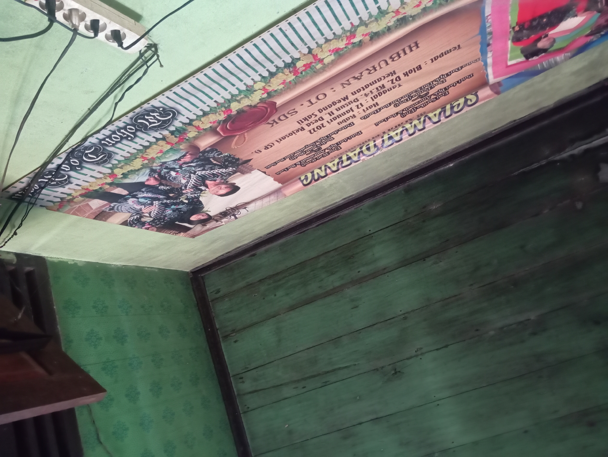 The image depicts an interior scene, likely a room in Rejosari, Indonesia. The focus is on a horizontal banner that is partially unfurled and attached to a wall. The banner is adorned with a floral pattern and features text in Indonesian. The text indicates details about an event, including "HIBURAN : OT. SDK", "Tempat: Blok D2 RT.14, Kecamatan Mesjid, Desa Rejosari (SP )", and "Tanggal : Hari: Rabu, Tanggal: 12 Januari 2023". The banner also shows photographs of people, possibly participants or performers at the event. The surrounding environment includes a green-patterned wall and a wooden floor with visible planks. Electrical cords are draped across the banner and a power strip is mounted on the wall. The lighting appears to be from an overhead source, casting shadows and highlighting the textures of the surfaces. The overall impression is one of an ordinary domestic or community space where an event is being advertised or documented.