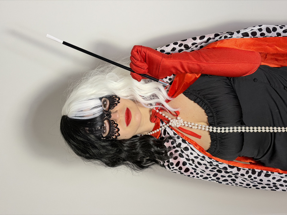 The image features a single individual, appearing to be an adult female, dressed in a costume against a plain, light-colored wall, indicating an indoor setting. The individual wears a bicolored wig, with black hair on the viewer's left side and white hair on the viewer's right side, complete with bangs. A black lace masquerade mask covers the eyes and upper nose, and red lipstick is visible on the mouth. She is adorned with a long pearl necklace, featuring a decorative clasp, draped around her neck and down her torso. Her costume includes a black top or dress with a low neckline and a white cape with black dalmatian-like spots, lined with vibrant red fabric, tied at the neck with red ribbons. Long red gloves cover both forearms. In her gloved right hand, she holds a slender black cigarette holder with a white tip, positioned near her chest. The individual faces forward, directly towards the viewer. No specific geographical landmarks identifying the location within Budva, Montenegro, are visible in the background.