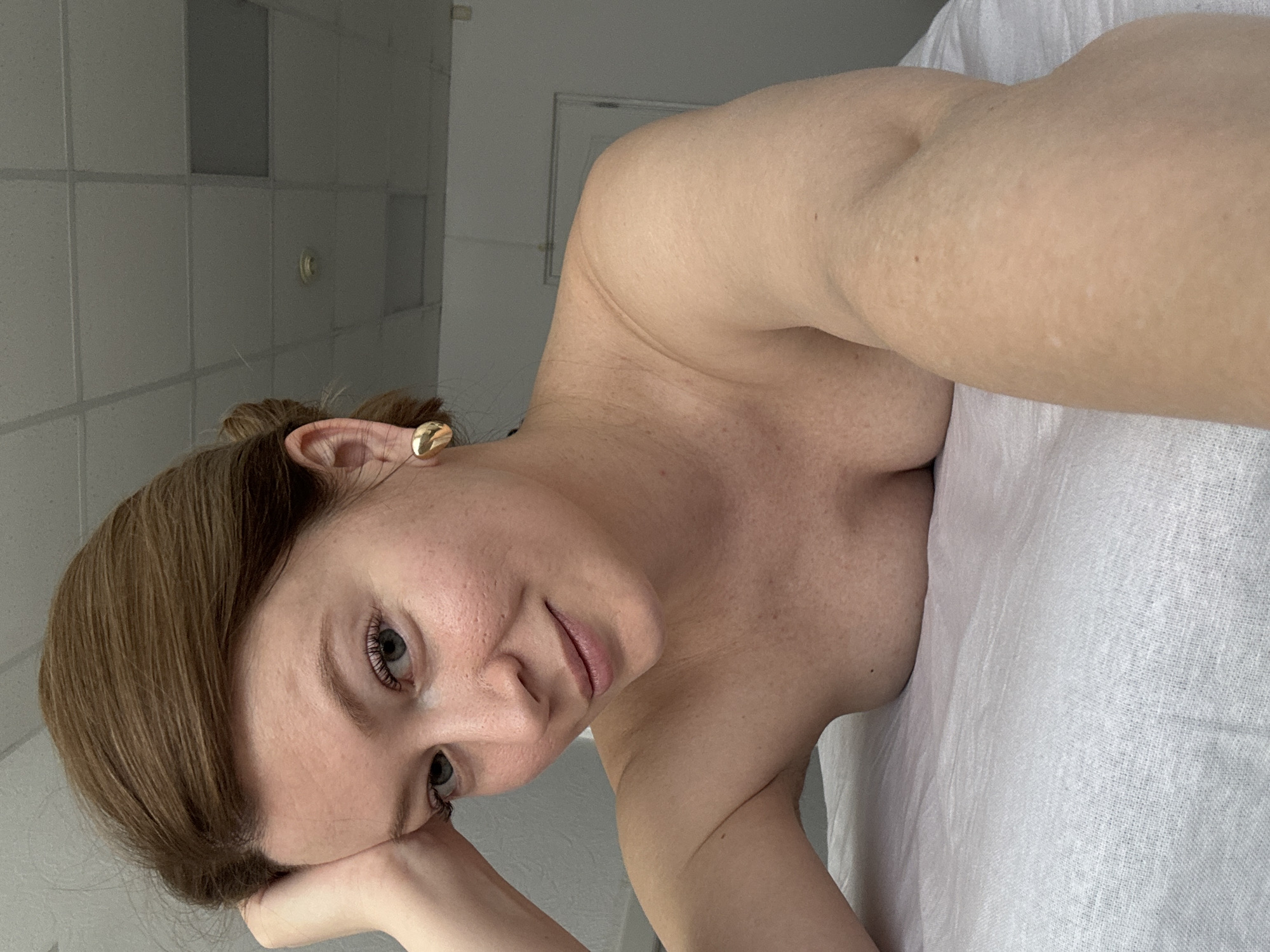 A woman with brown hair is lying on a white surface, possibly a bed or examination table. She is wearing a gold earring in her left ear. The background shows a tiled wall and ceiling. The image location within Chaikovsky, Russia cannot be determined from the visual information.