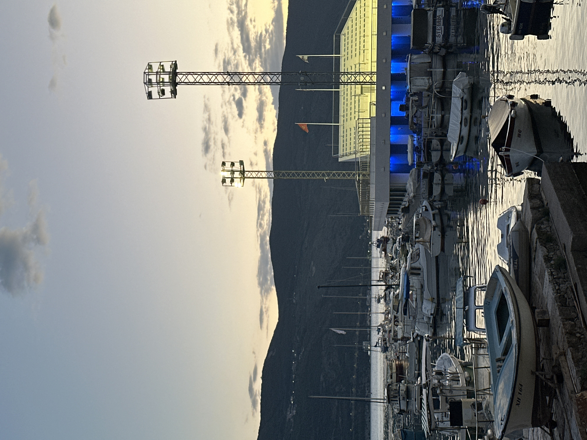 The image depicts a harbor in Herceg Novi, Montenegro. A line of docked boats occupies the right side of the frame, situated beside a stone dock. Adjacent to the boats, a building with blue lighting extends along the harbor front. Two tall light towers stand behind the building, with a mountain range forming a backdrop. The sky above exhibits a mix of daylight and clouds.