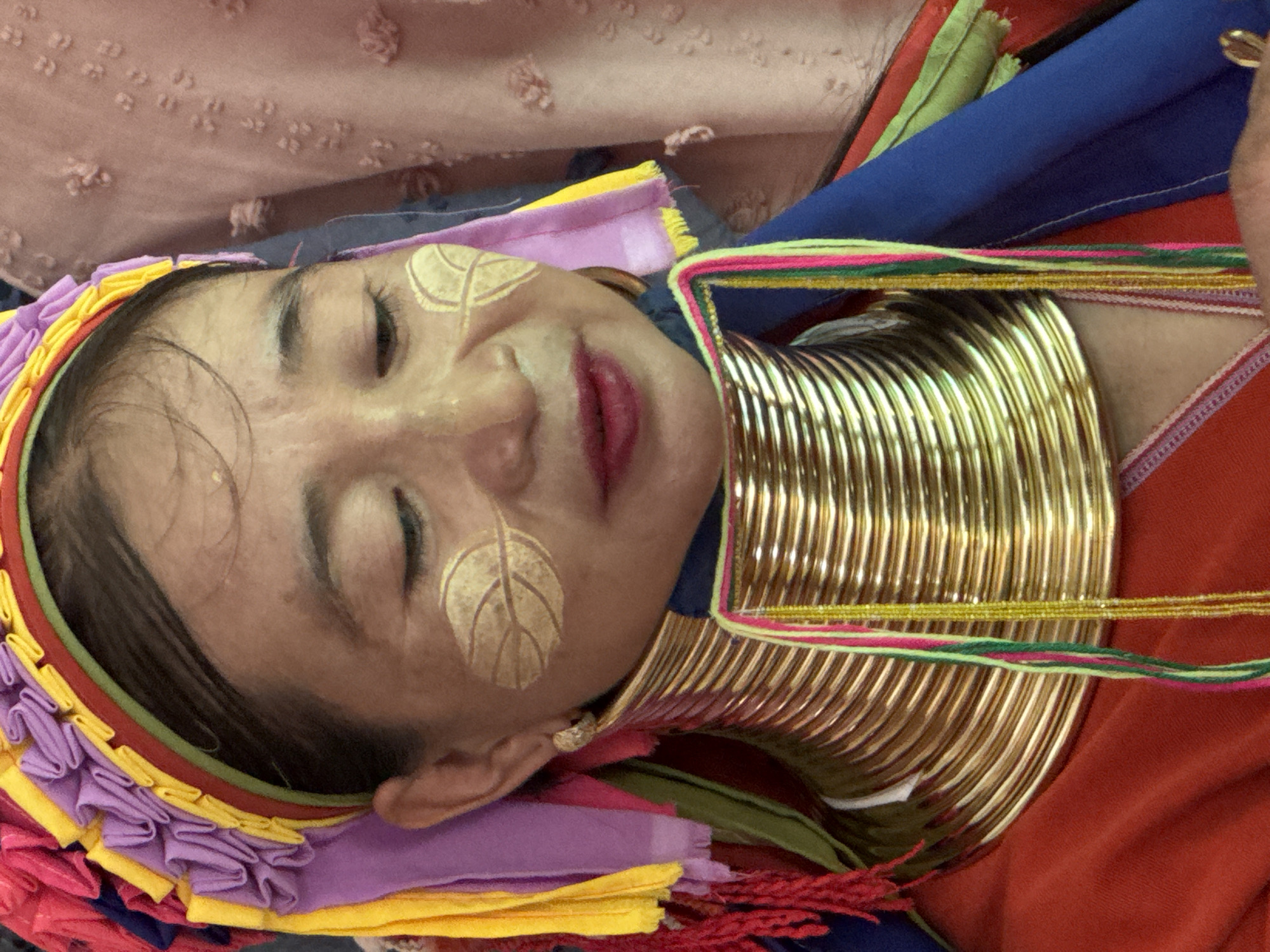 A woman with gold leaf designs on her cheeks lies with eyes closed wearing traditional neck rings. She has a colorful headpiece and is lying on a bed or cushion with various colors. The photo may have been taken in Ban Khlong Khian, Thailand.