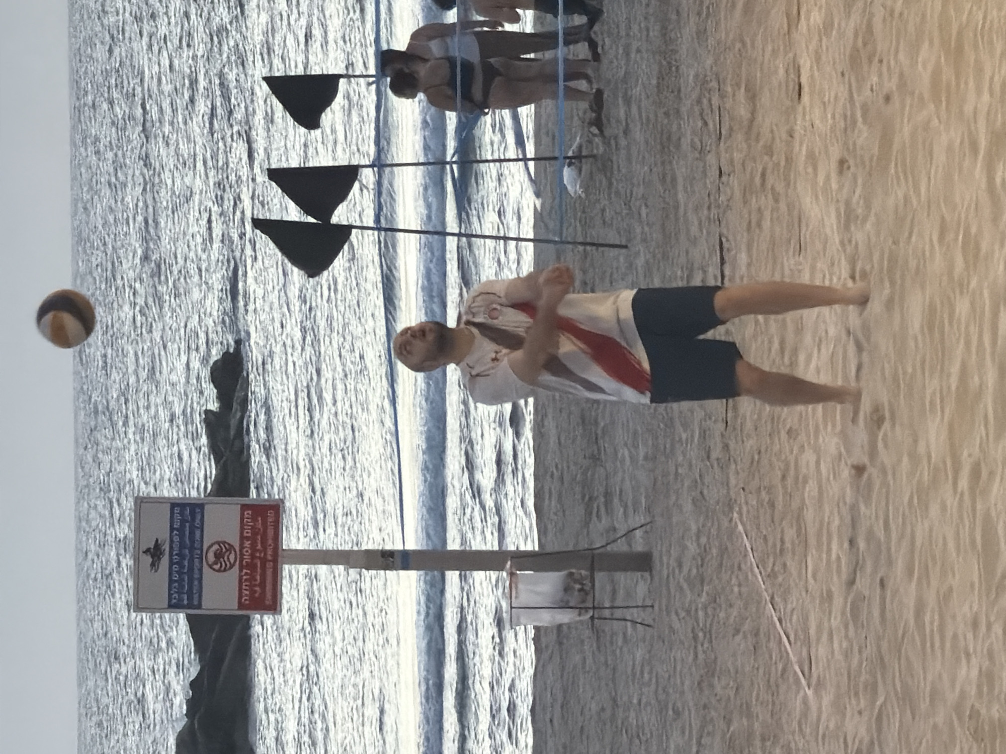 The image captures a beach scene in Tel Aviv, Israel. A man is captured in mid-motion, playing beach volleyball. In the background, others are gathered near the volleyball net. To the left, a signpost with dual-language signage (Hebrew and English) indicates designated areas for water sports and prohibits swimming. The Black flags nearby are posted because of dangerous water quality. The sea extends into the distance, and the beach itself takes up a significant portion of the foreground.