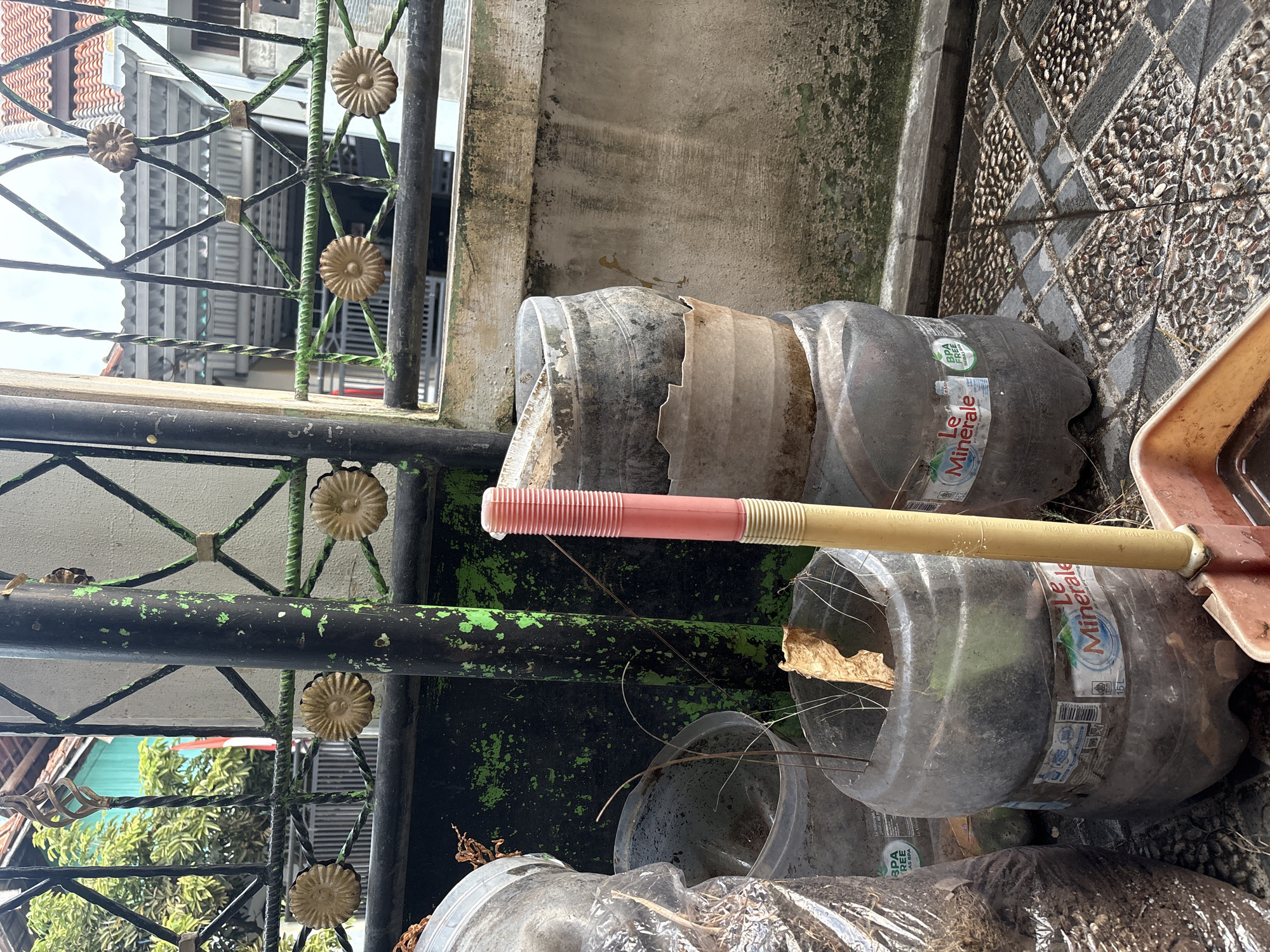 The image depicts an outdoor scene, likely a backyard or front yard, in Palimanan, Indonesia. A black metal fence with decorative gold floral accents runs across the background, partially obscuring a building with a tiled roof and a turquoise wall. In the foreground, several large plastic bottles, some appearing to be cut in half and repurposed as planters, are arranged on a paved surface. One bottle, a "Le Minerale" water bottle, is visible and appears to contain soil and dried leaves. A pink and yellow tool, possibly a broom or mop handle, leans against one of the bottles. The ground is tiled with a pattern of grey and brown stones. The overall impression is one of a somewhat cluttered outdoor space, potentially used for gardening or storage.