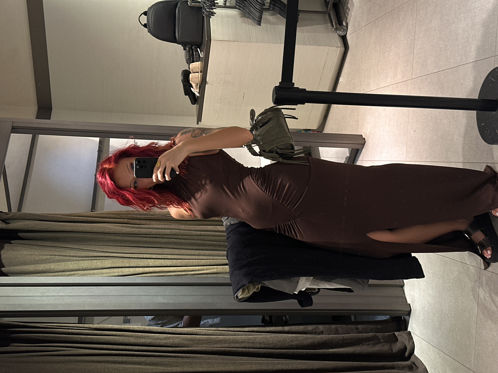 A woman with red hair is taking a selfie in a dressing room. She is wearing a brown dress and black sandals. The dressing room has beige curtains and a gray frame. Clothes are hanging on a rack in the background. The location is likely a clothing store, and the image does not confirm the location is Kuta, Indonesia.