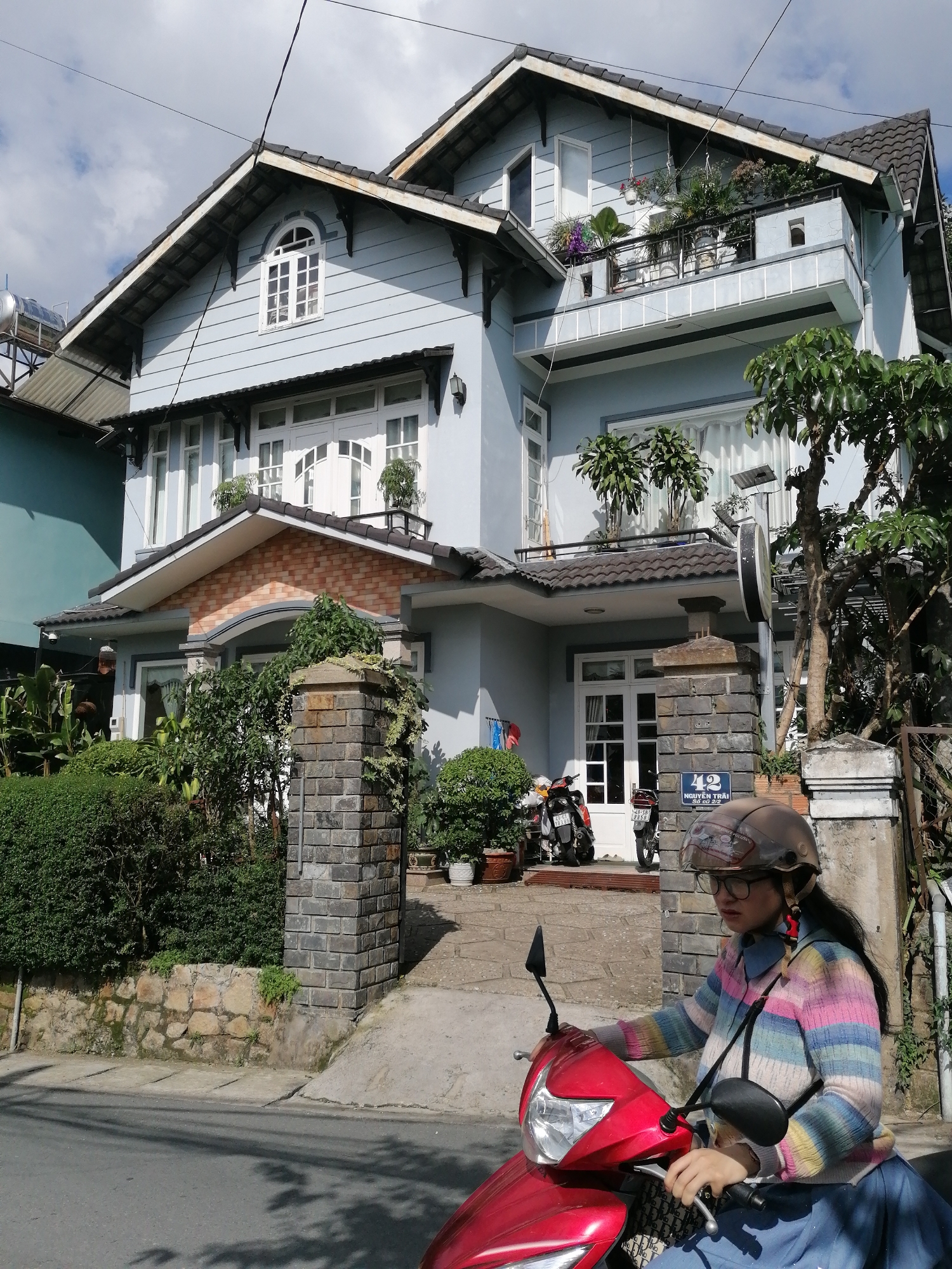 This image captures a street scene in Phường Xuân Hương, Đà Lạt, Vietnam, on a bright, partly cloudy day. A light blue, two-story house with a dark tiled roof dominates the background. The house features several windows, including a distinctive arched window on the upper level, and a balcony with potted plants. The exterior walls are clad in horizontal siding.
In the foreground, a young woman wearing glasses and a colorful striped sweater is riding a red scooter. She is wearing a brown helmet. The scooter has a black rearview mirror. To the left of the scooter, two motorcycles are parked.
The street is paved, and there is a sidewalk with a low stone wall and dense green hedges. A stone pillar, likely part of a gate or fence, stands to the right of the woman, with a sign on it displaying the number "42" and Vietnamese text. Trees with green foliage are visible around the property. The overall impression is of a residential area in a slightly hilly urban landscape.