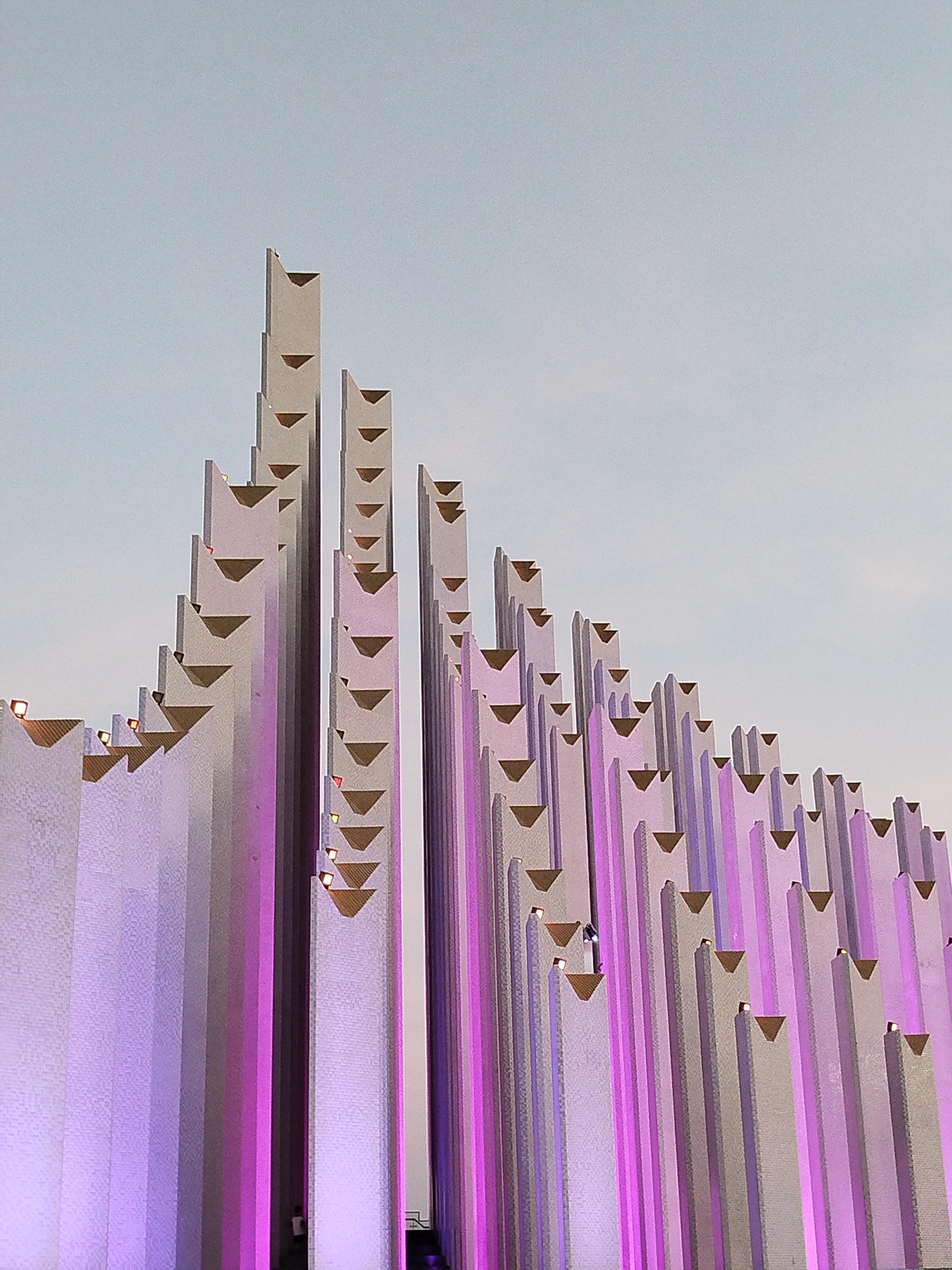 The image shows the Vietnam National University, Ho Chi Minh City, Vietnam, a monument consisting of numerous tall, gray concrete pillars of varying heights. The pillars are arranged in a rising pattern, tallest on the left and gradually decreasing in height to the right. Each pillar has an inverted triangular recess near the top. Some of the recesses contain small light fixtures, illuminated in warm yellow. The pillars are lit with a diffuse pink or purple light washing up from the base. The sky is light blue and slightly overcast. There is a small figure visible at the base of the pillars on the left, and another climbing the pillars.