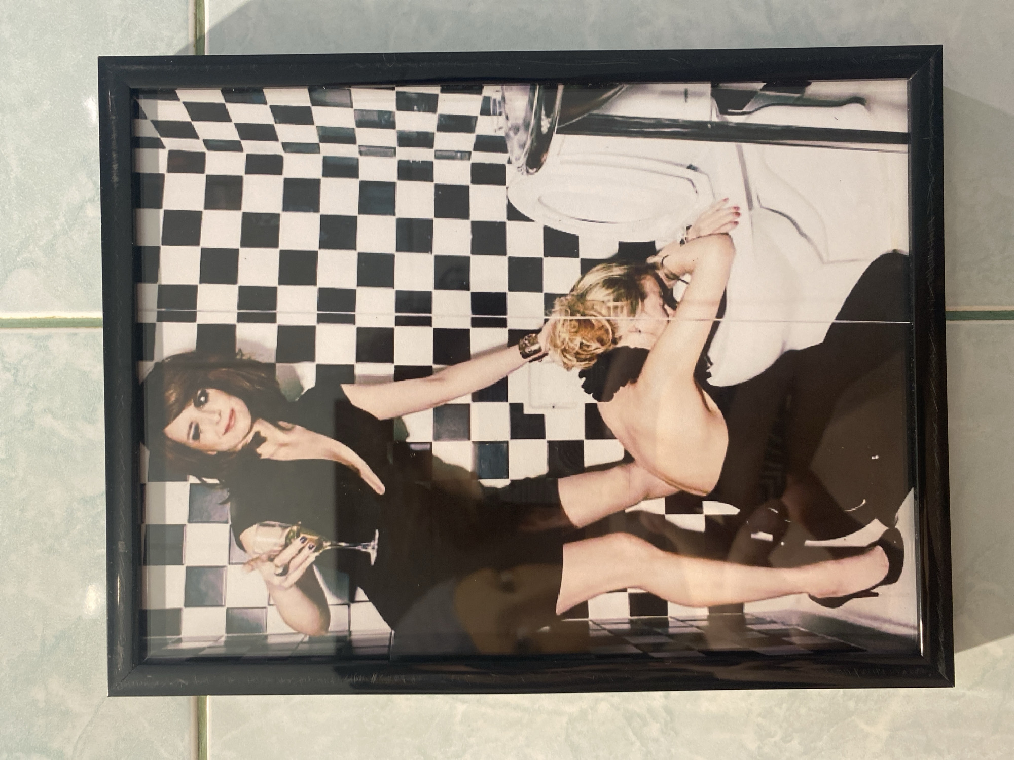 The image is a framed photograph. The photograph features two women in a bathroom with a black and white checkered floor. One woman, with dark hair, is holding a champagne flute and looking at the viewer. The other woman, with blonde hair, is leaning over a toilet. The location appears to be a bathroom, but no city information is present.