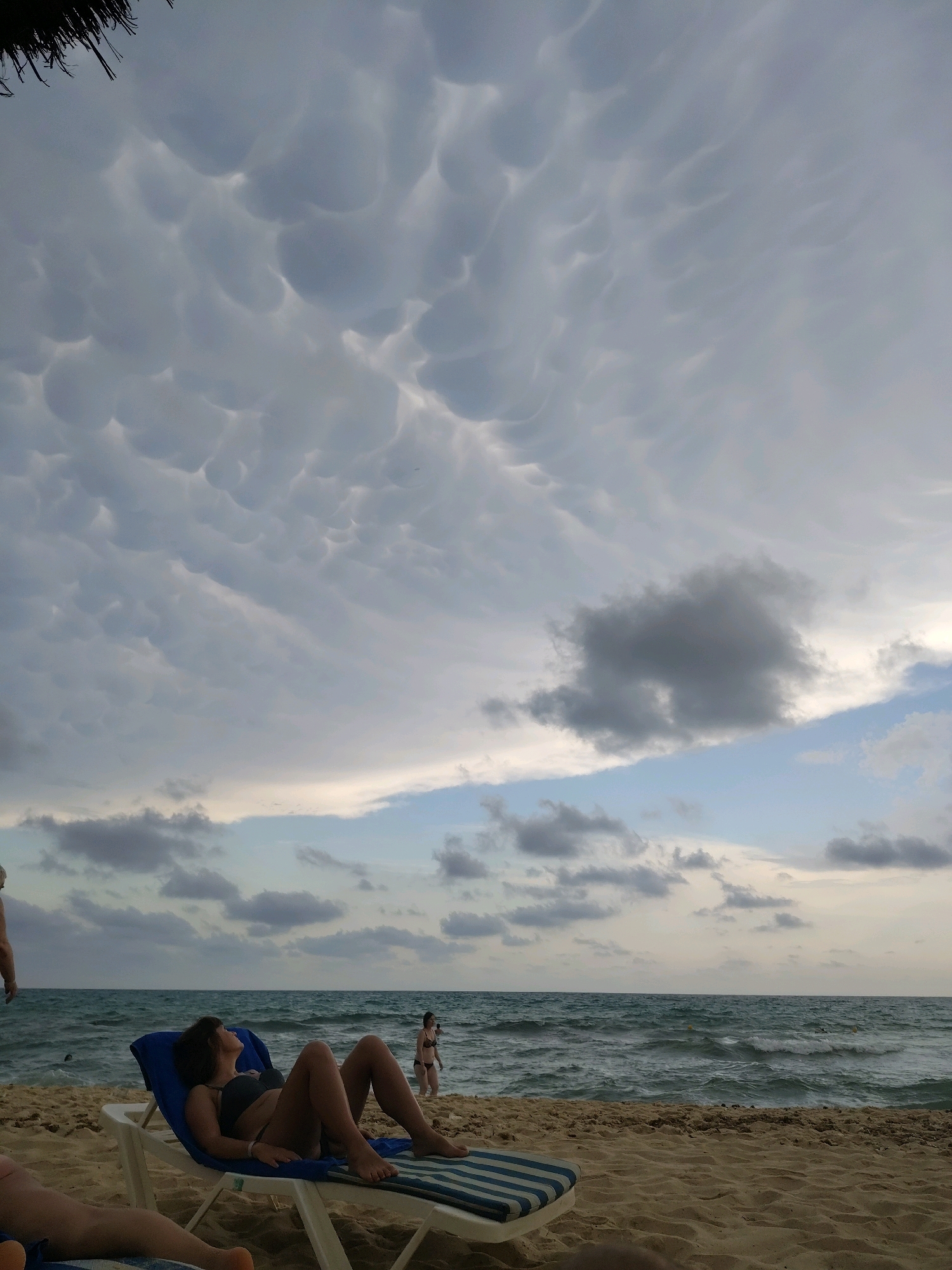 This is a wide shot of a beach in Yasmine Hammamet, Tunisia, featuring a dramatic cloudy sky over the ocean. In the foreground, a woman is reclining on a blue lounge chair on the sand. She is wearing a dark bikini and looking upwards at the sky. To her left, another person's legs are visible, resting on a striped lounge chair. Further back, near the water's edge, two more figures are present, one wading in the shallow water and the other standing. The ocean is choppy with waves rolling towards the sandy shore. The sky is filled with unusual, mammatus clouds, which appear as rounded pouches hanging from the underside of a cloud. A portion of a thatched roof structure is visible in the upper left corner of the image.