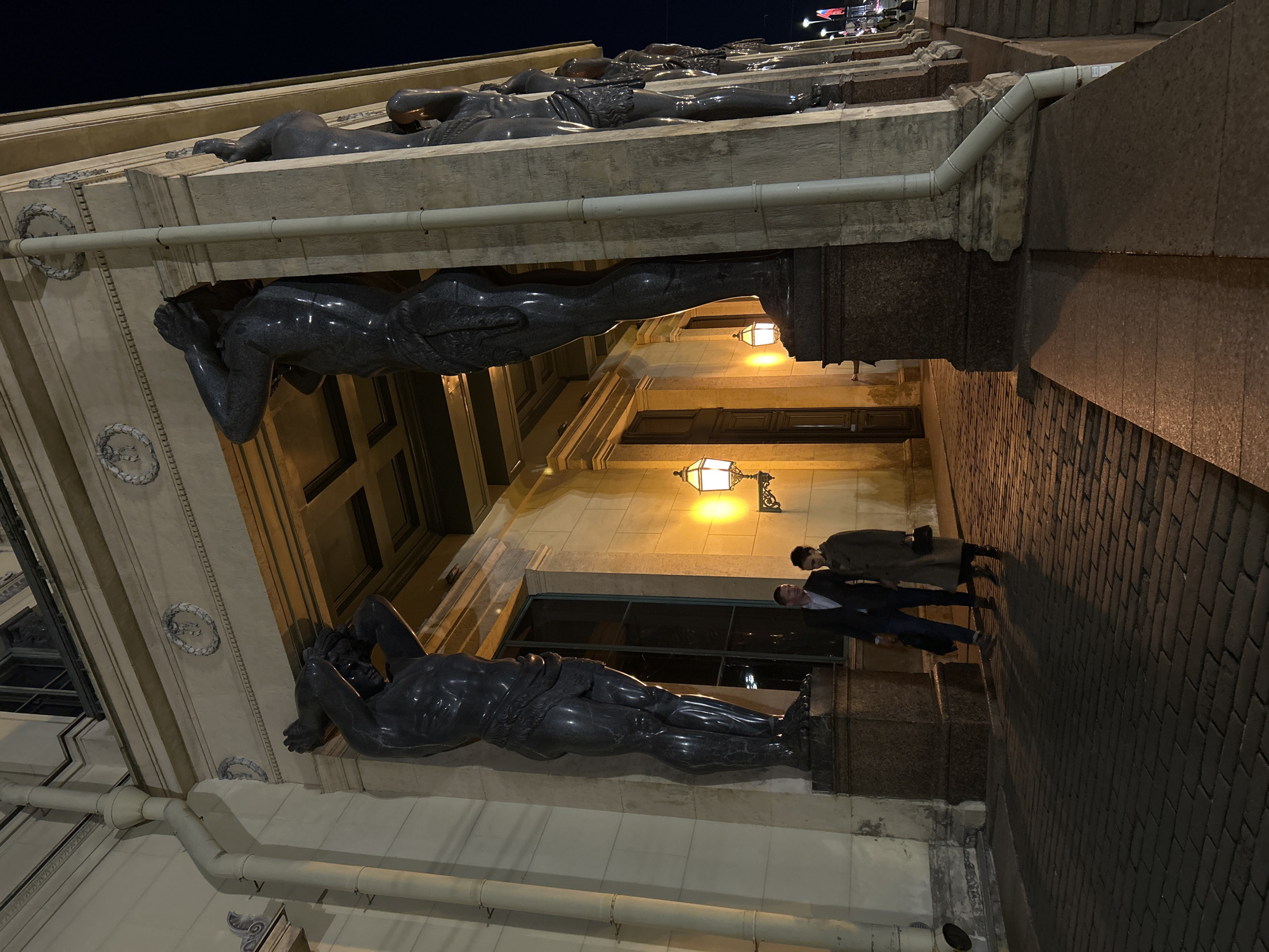 The image depicts a nighttime scene outside a building in Saint Petersburg, Russia. The building's facade is adorned with caryatids, sculptural figures of men that appear to support the structure above. Two people are walking on the sidewalk in front of the building. The street is paved with dark stone bricks. Street lamps illuminate the building's facade and the sidewalk. The sky is dark.