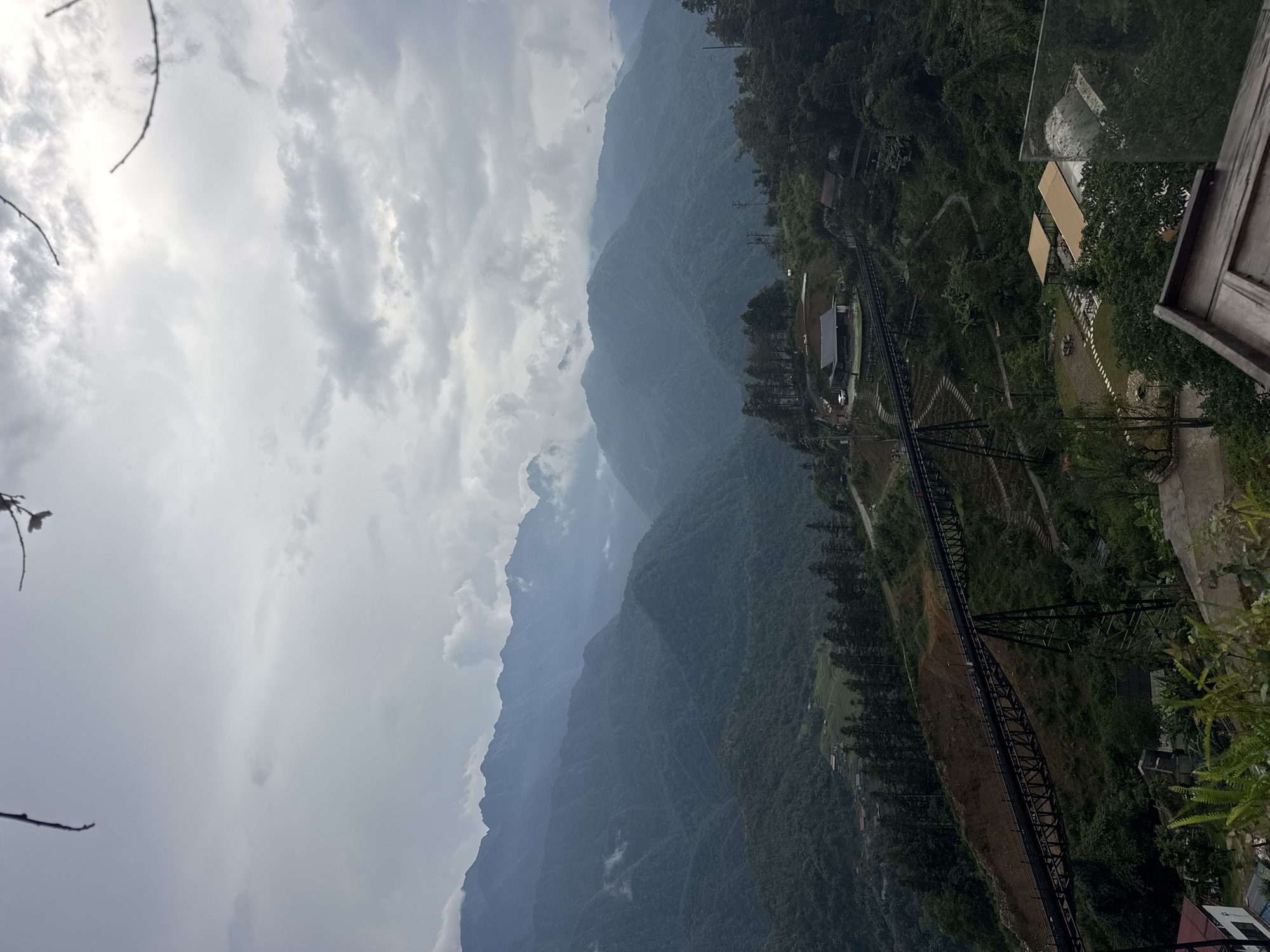 The image depicts a panoramic view of a mountainous landscape in Sa Pa Ward, Vietnam. The foreground features a modern building with a wooden structure and a reflective glass barrier, suggesting a viewpoint or restaurant. In the midground, a dark metal trestle bridge traverses a steep, vegetated slope, with a road winding alongside it. Below the bridge, several small buildings with light-colored roofs are nestled amidst trees. The background is dominated by lush green mountains, shrouded in a misty haze, with layered peaks receding into the distance under a cloudy sky. The overall scene conveys a sense of natural beauty and human infrastructure integrated into the rugged terrain.