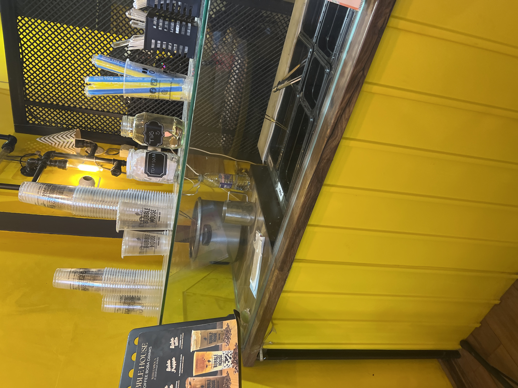 The image, rotated 90 degrees clockwise, depicts a vibrant counter area within a bubble tea shop, likely located in Plovdiv, Bulgaria. The backdrop is a bright yellow wall with horizontal paneling, providing a striking contrast to the counter's contents.
On a glass-topped counter, various items for drink preparation are arranged. To the left, a black mesh rack holds clear utensils. Below it, stacks of clear plastic cups, many branded with "BUBBLE HOUSE ORIGINAL TASTE", are visible. Blue and yellow straws, some with "PULL HERE" text on their wrappers, are stored upright in a container. Adjacent are two glass jars: one filled with coins and labeled "TIPS", and another marked "TO M 50 ML". Light fixtures and a water bottle are also present on the lower counter surface.
Towards the right, a metal shaker or measuring cup sits next to a section containing multiple black rectangular inserts, possibly for holding ingredients or ice, with small tongs resting inside. The counter edge is finished with a dark wooden trim. A partially visible menu board in the bottom left corner advertises "BUBBLE HOUSE COFFEE BOBA DRINKS WITH TAPIOCA", showcasing options like "Boba Frappe" and "Boba Latte". No people are visible, suggesting a moment of inactivity or preparation.