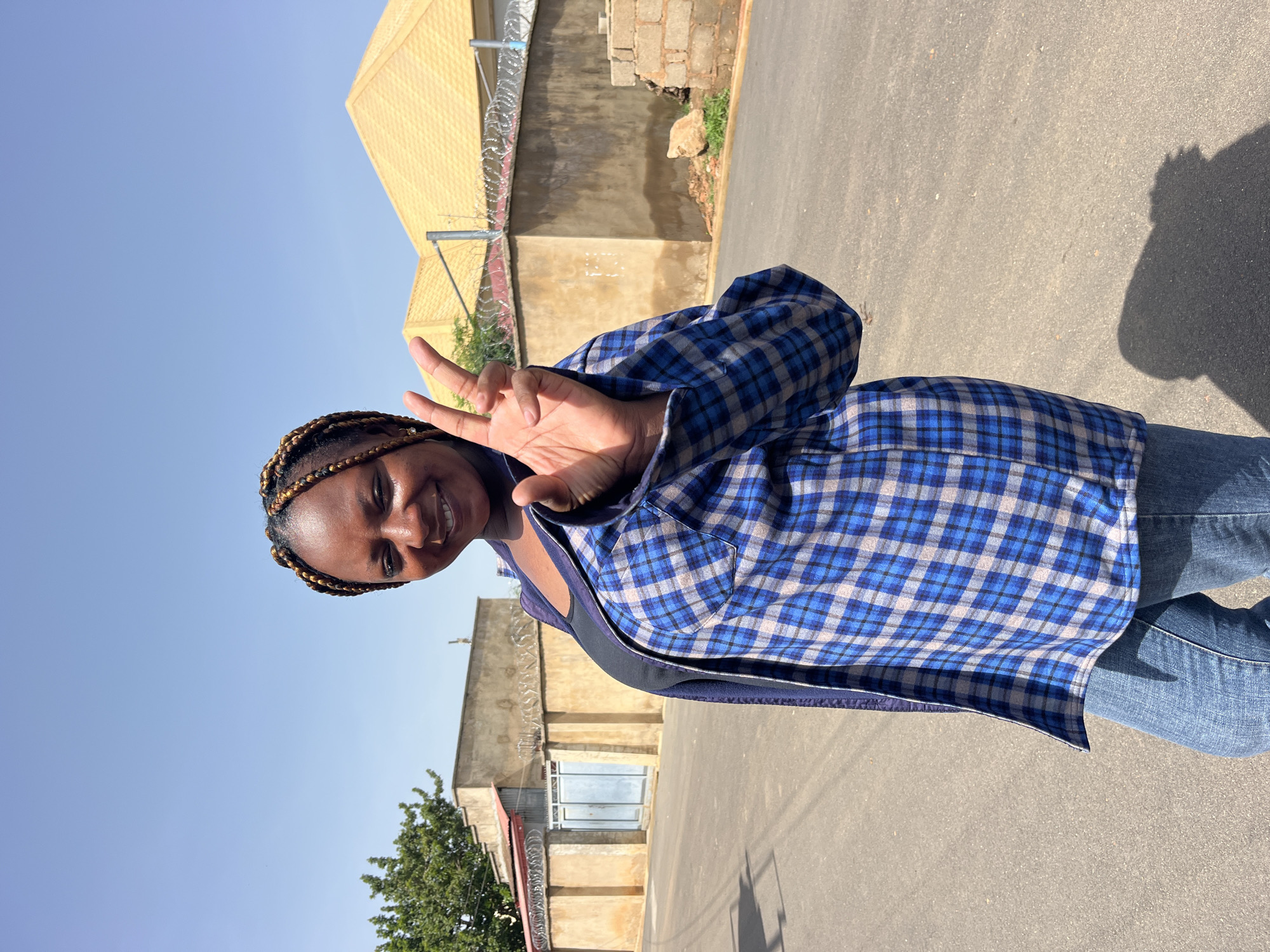 A medium shot captures a Black woman with braided hair standing outdoors on an asphalt road. She is smiling and holding up her right hand with two fingers extended in a peace sign. She is wearing a blue and white plaid shirt over a dark top and blue jeans. In the background, a beige wall with barbed wire on top runs along the side of the road, with a yellow-roofed building behind it. The sky is clear and blue.
