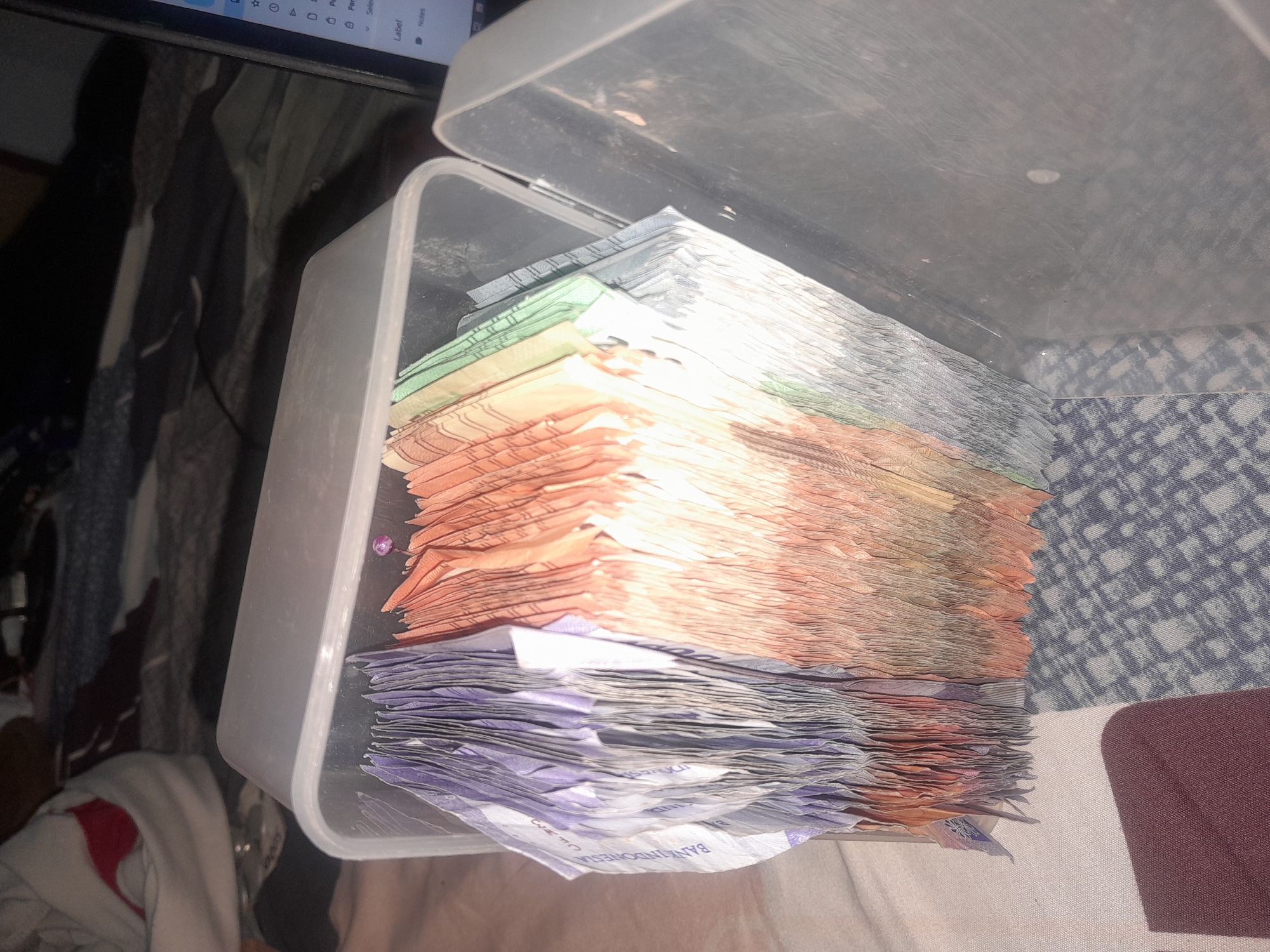 A clear plastic container filled with stacks of Indonesian Rupiah banknotes is placed on a patterned fabric surface. The banknotes are neatly fanned out, revealing various colors including purple, orange, green, and blue, suggesting different denominations. Some of the banknotes have text visible, including "BANK INDONESIA". The container appears to be in a domestic setting, with a blurred background showing what could be a bed or sofa and a laptop. The lighting is dim, casting shadows on the fabric. The image does not contain information about the specific location in Keboharan, Indonesia.
